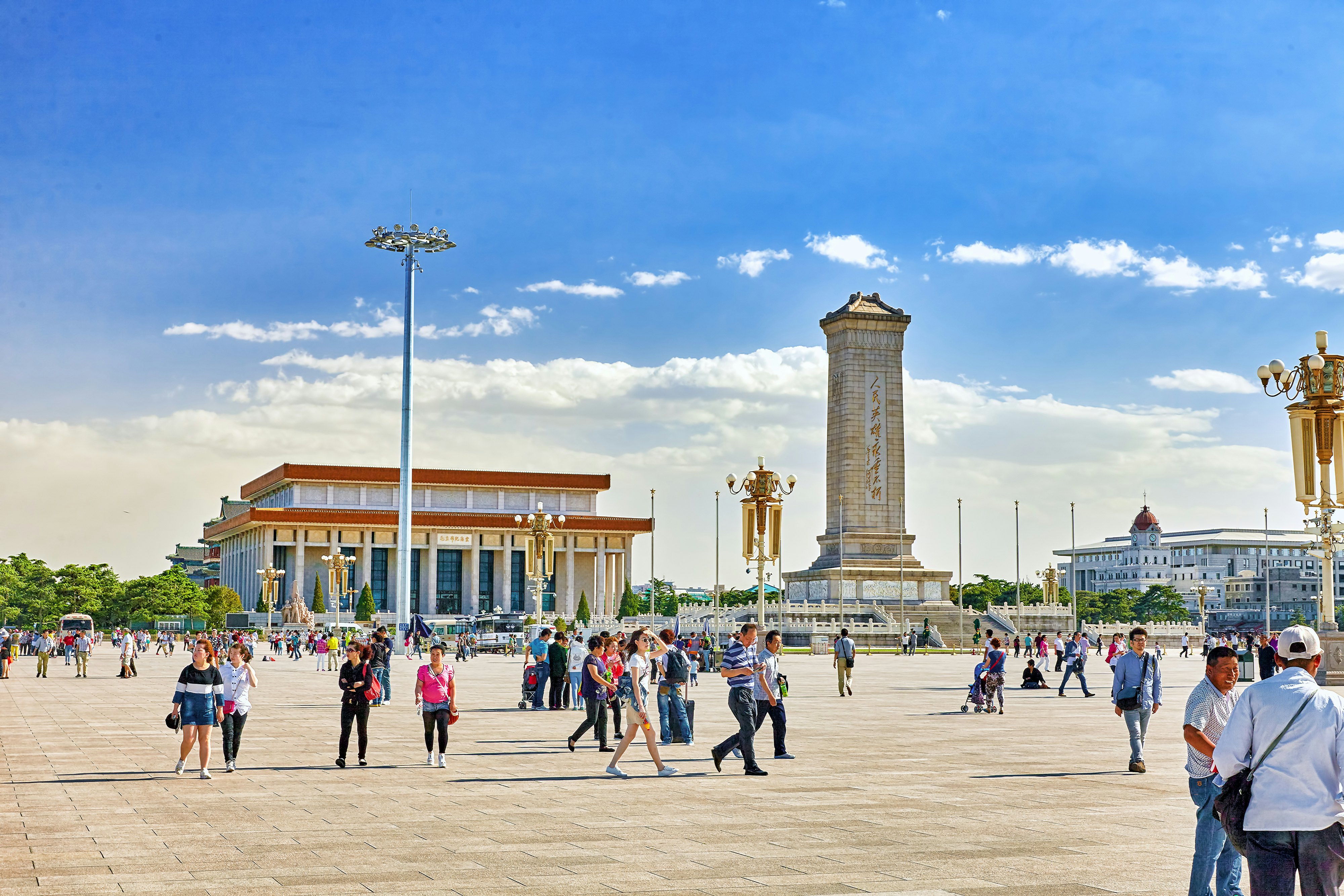 People walk through a large city square lined with large buildings and a tall stone tower.