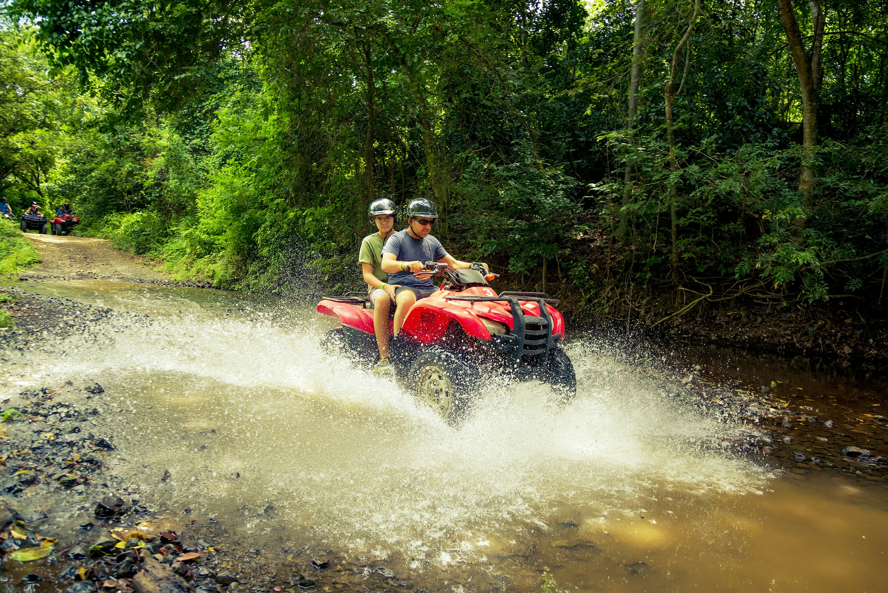 Two people drive a red 4WD vehicle through a large muddy puddle on a dirt track through a forested area in Costa Rica.