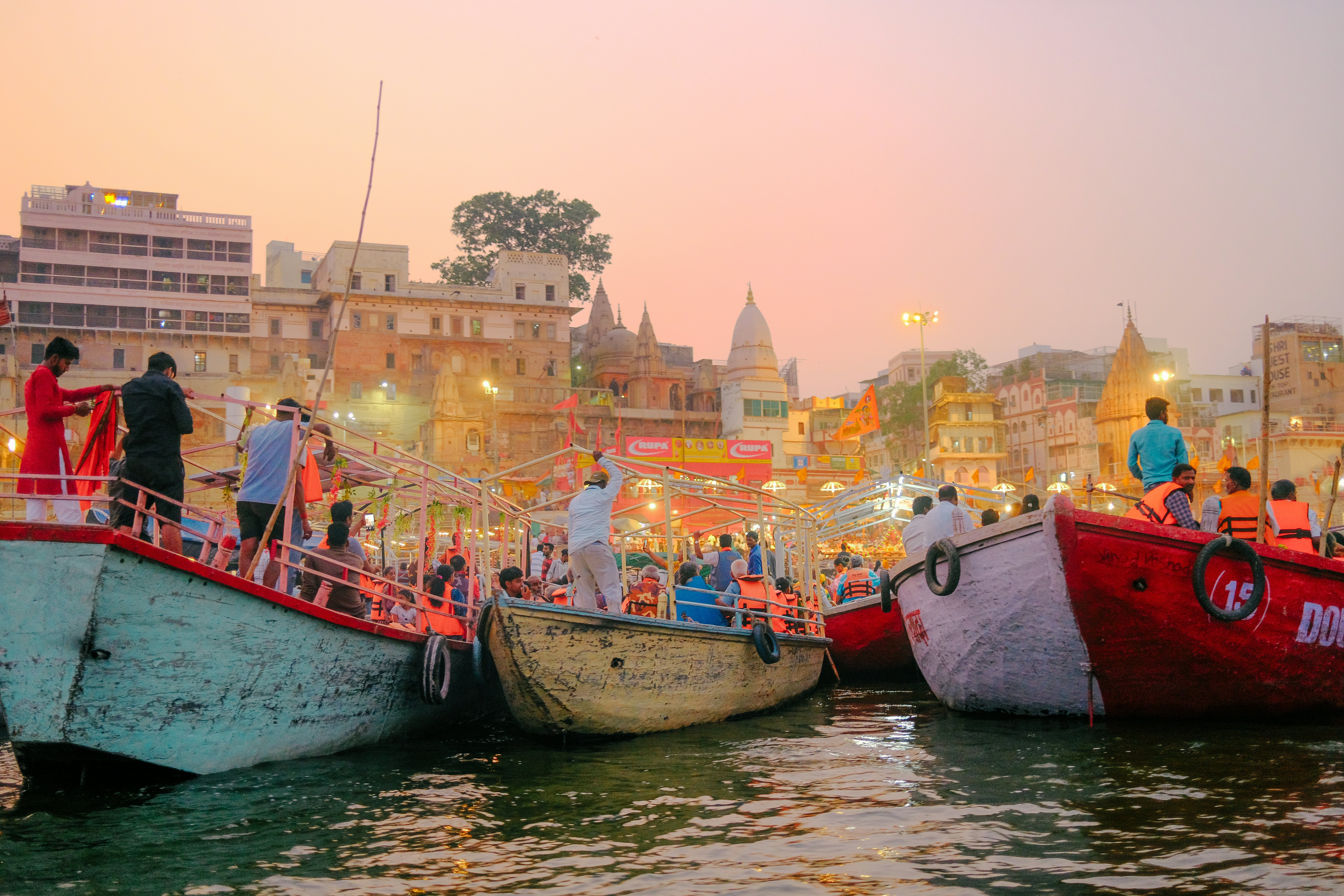 A river packed with small boats that are loaded with people on the edge of a city where the twilight sky is glowing pink.