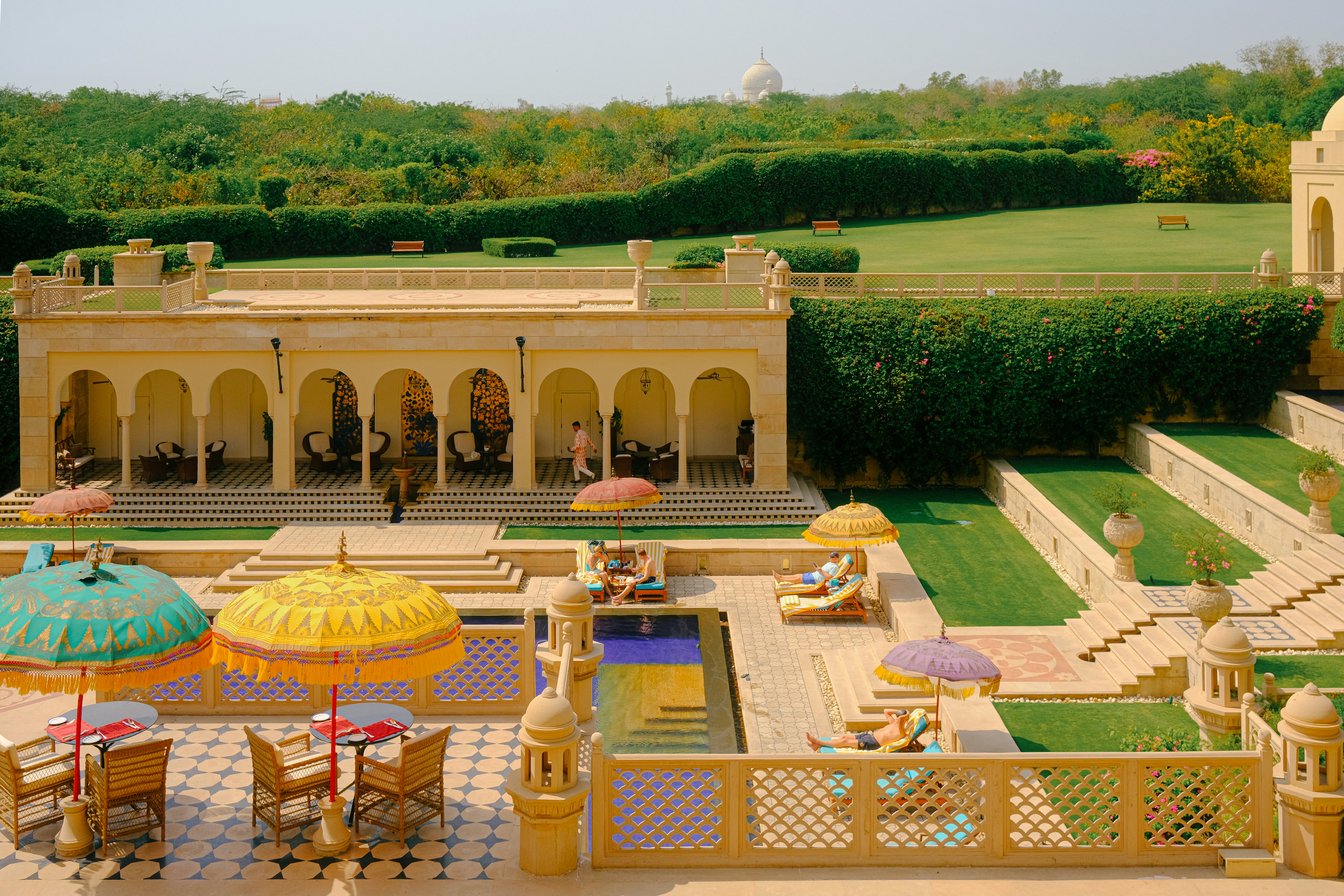 The poolside terrace of a luxury hotel with brightly colored umbrellas providing shade to the guests. A large white marble dome peeks over the gardens in the distance.