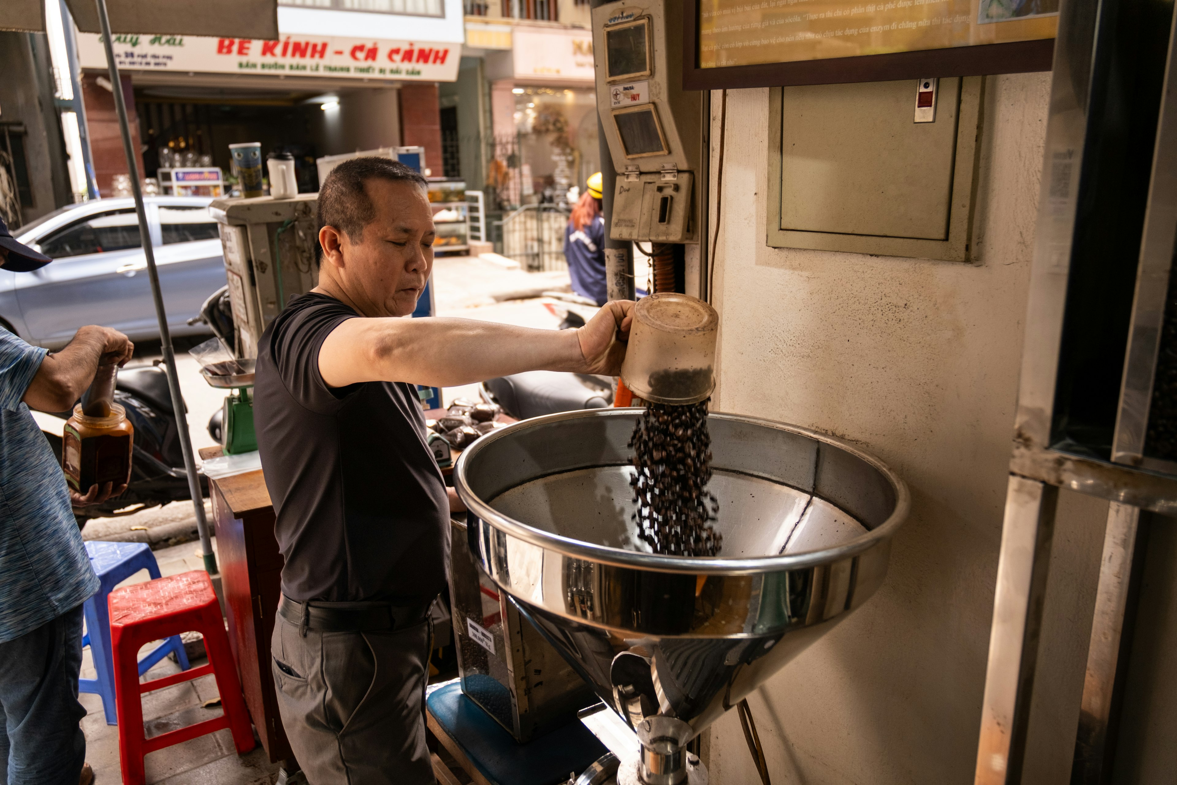 A man empties a bucket of coffee beans into a large grinder at a cafe