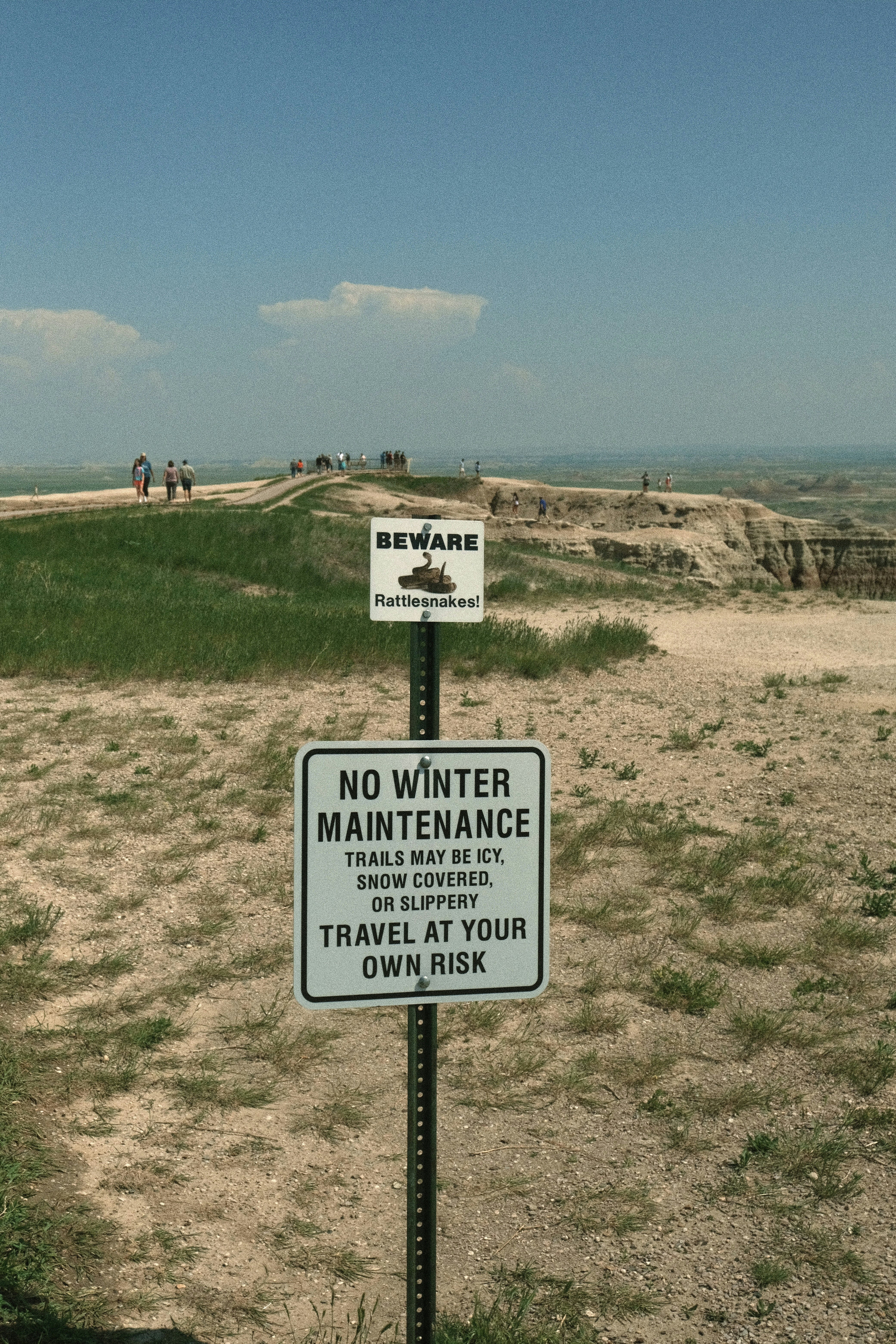 "Beware rattlesnakes" sign in grasslands near a path with people walking on it.