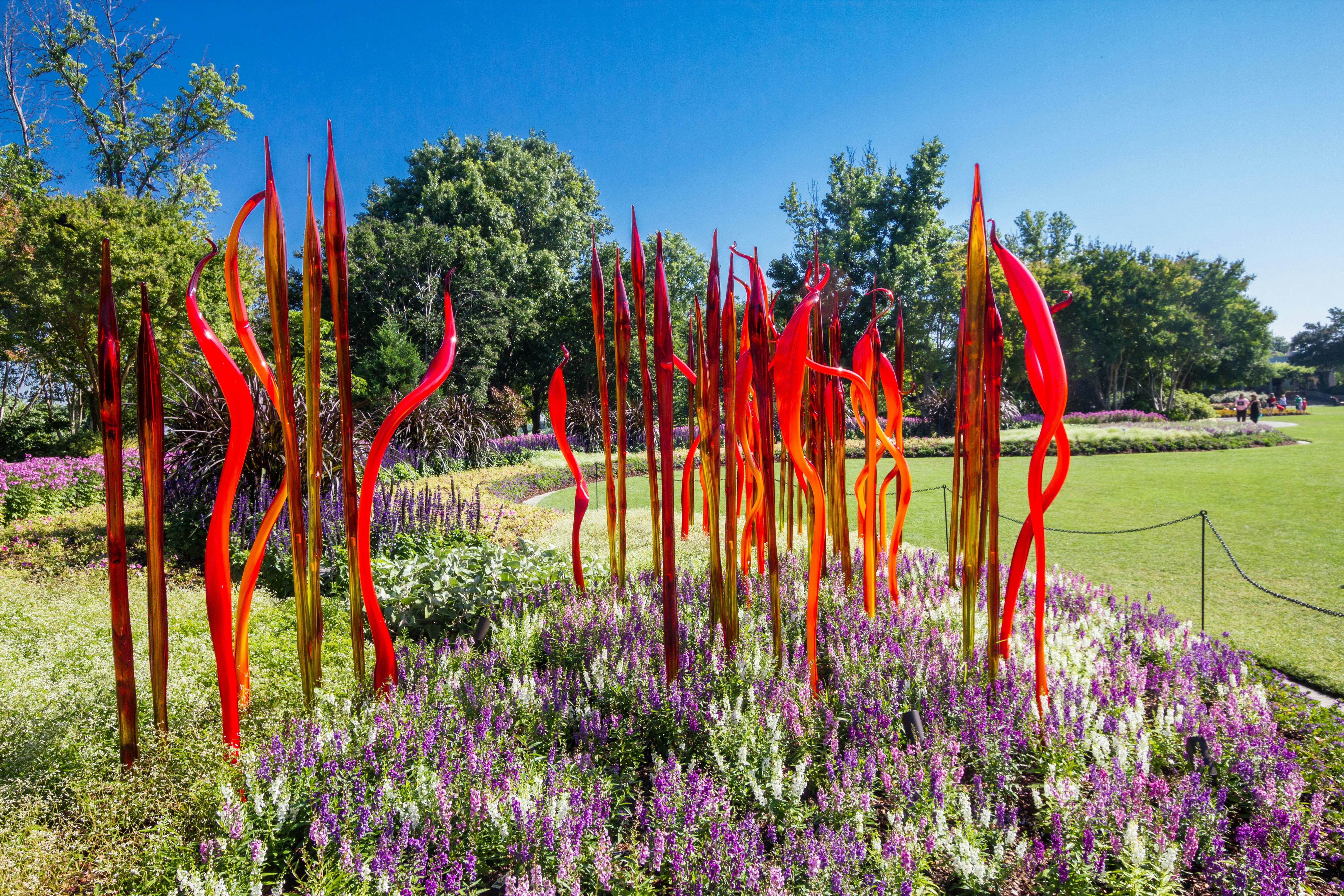 Tall, narrow red glass sculptures set in a garden of purple flowers with grass and trees beyond on a sunny day.