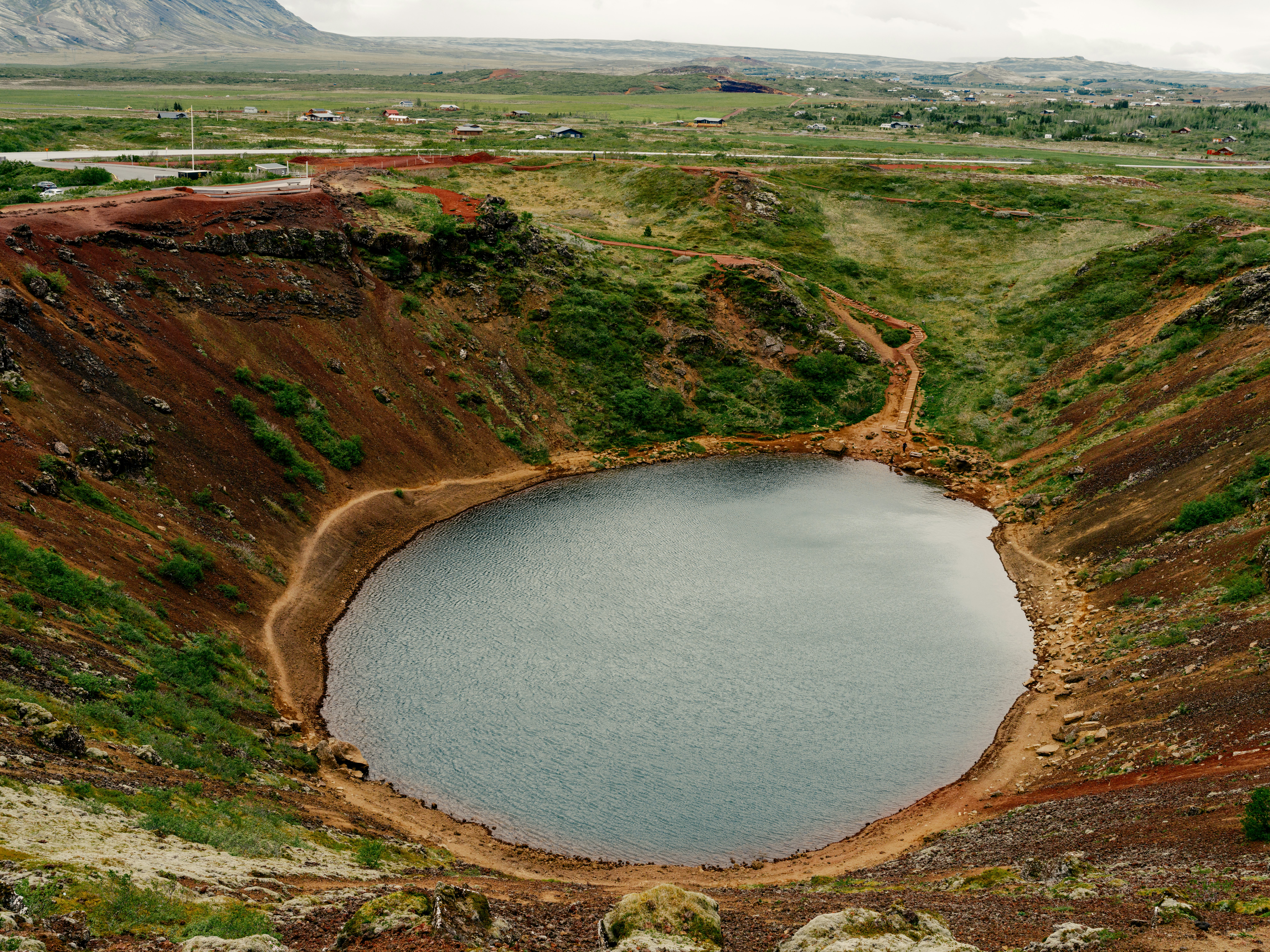 Large water-filled crater surrounded by grass- and dirt-covered hillside on an overcast day.