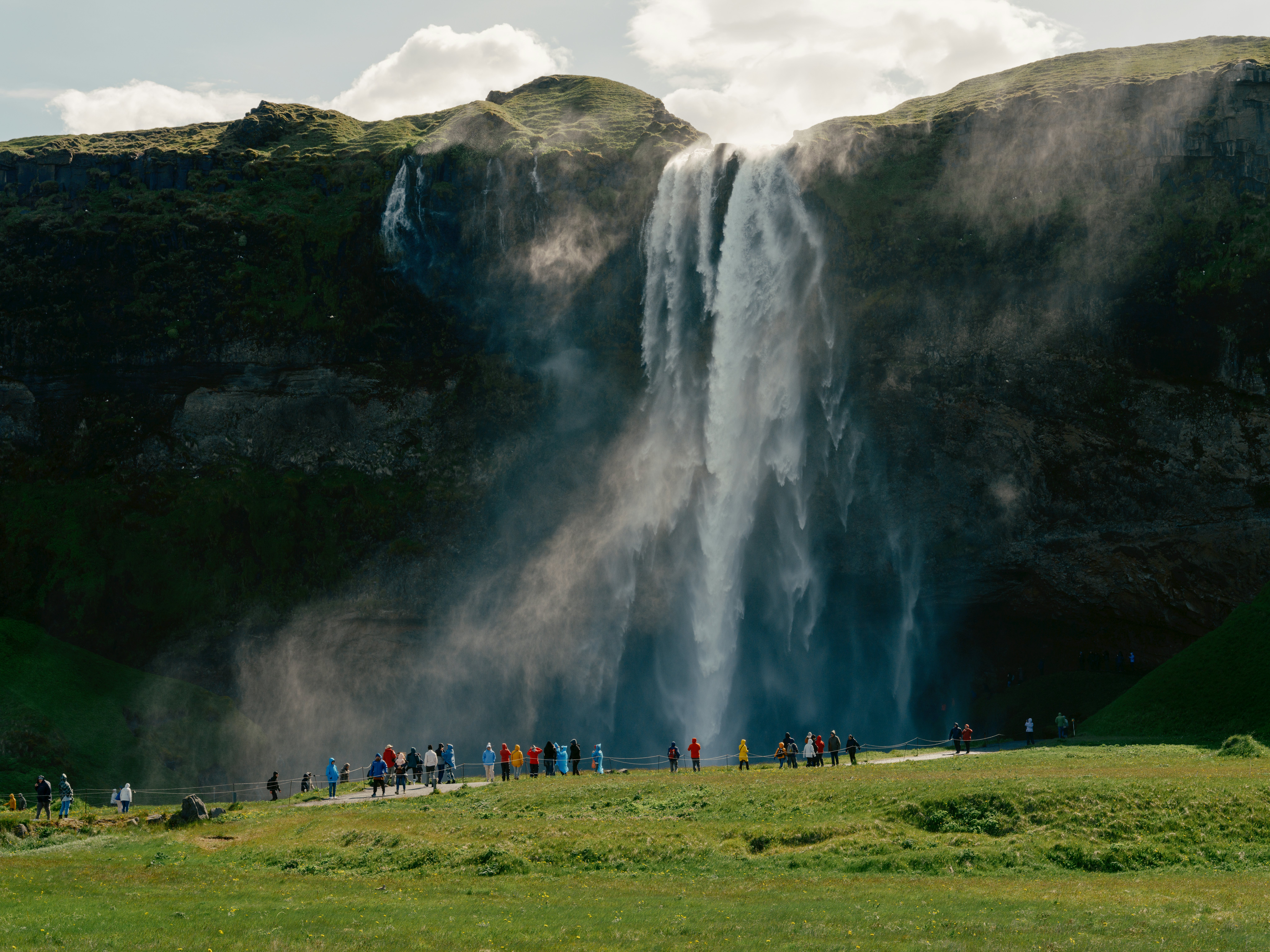 People in multi-colored jackets along a walkway with grass in the foreground and a high waterfall in the distance on a mostly sunny day.