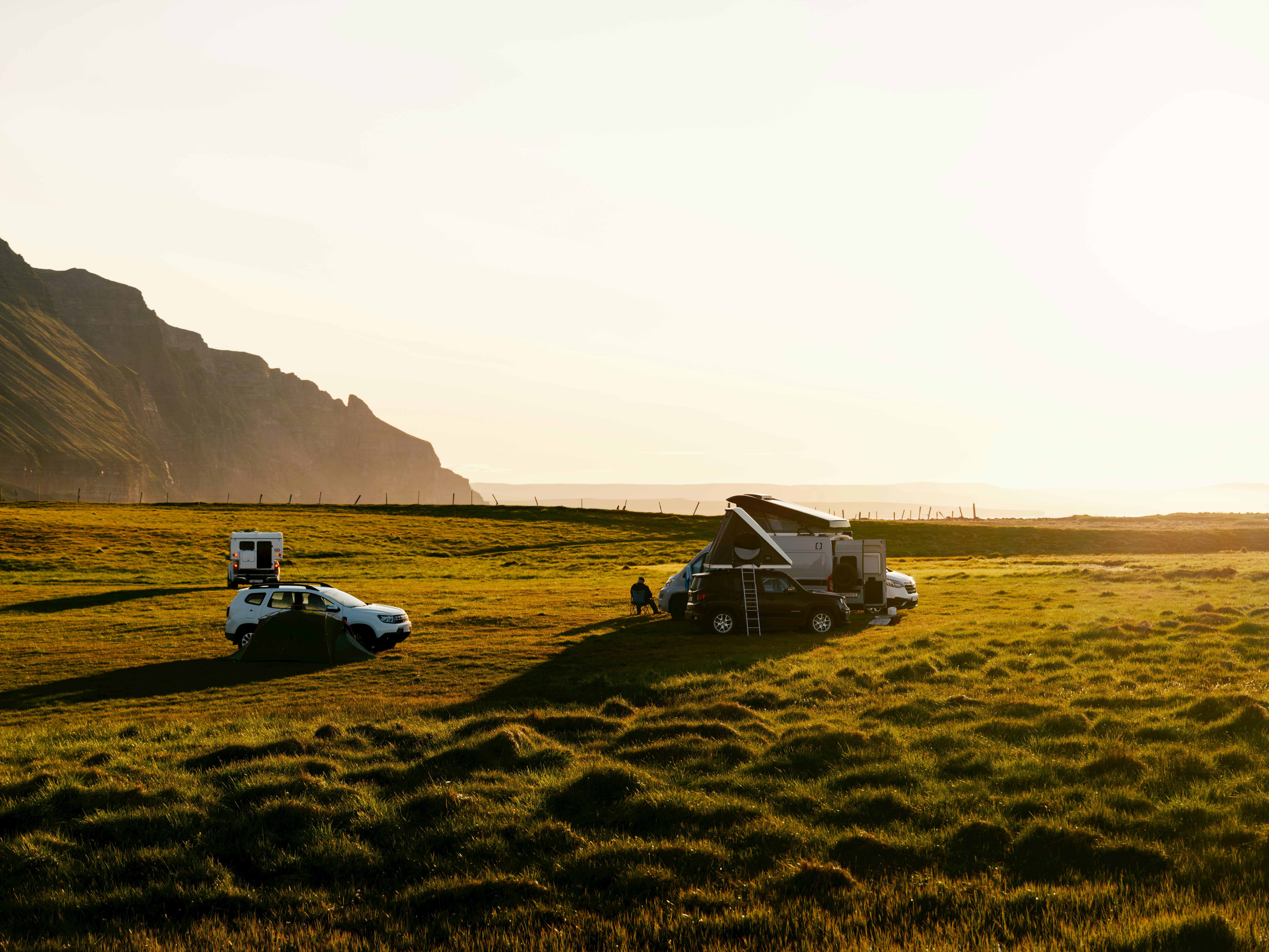Two recreational vehicle camping vans and two SUVs parked in a grassy field with a rocky hillside in the distance with bright sun late in the day.