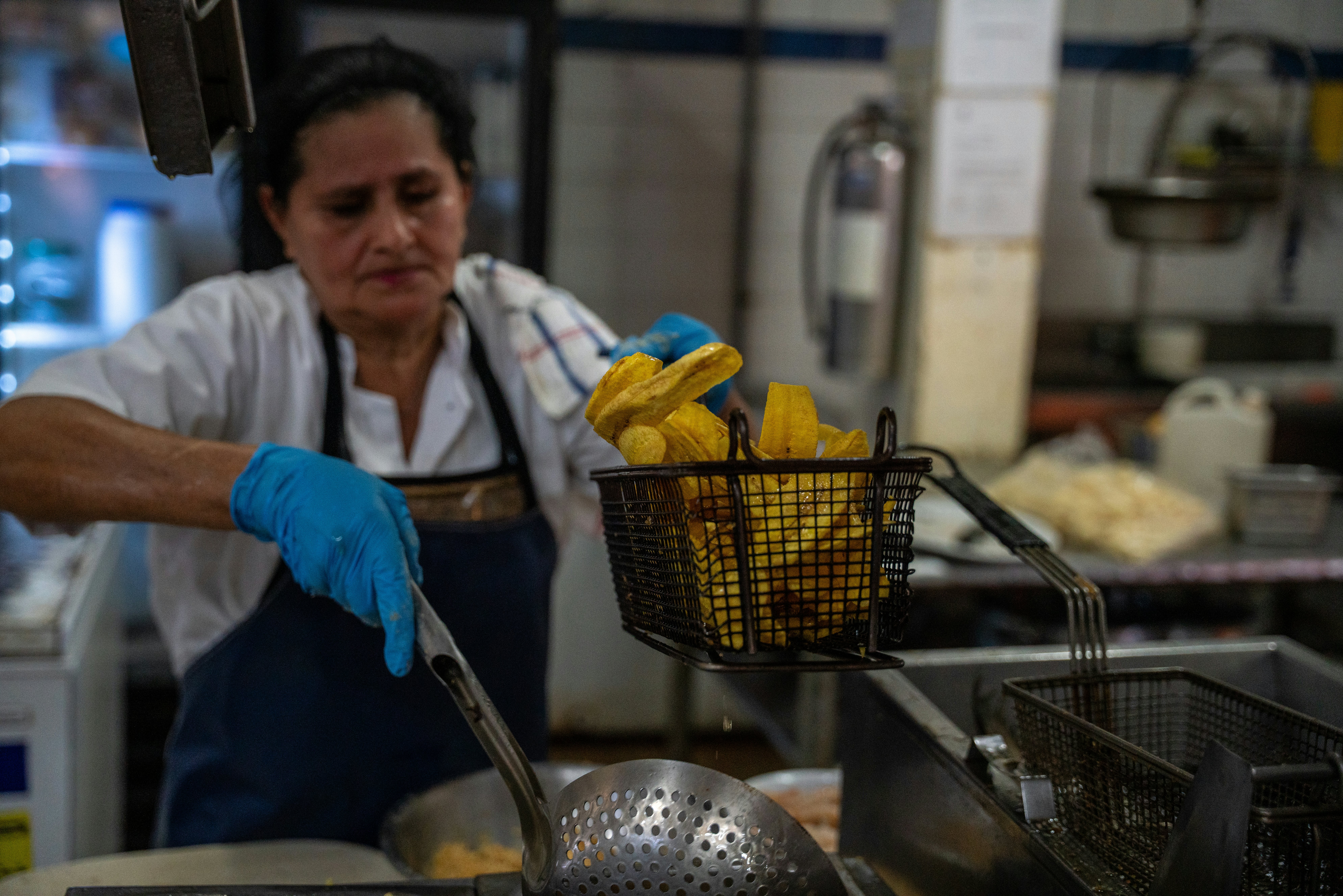 A woman in an apron and gloves holds a basket of fried plantains above a strainer in a commercial kitchen.