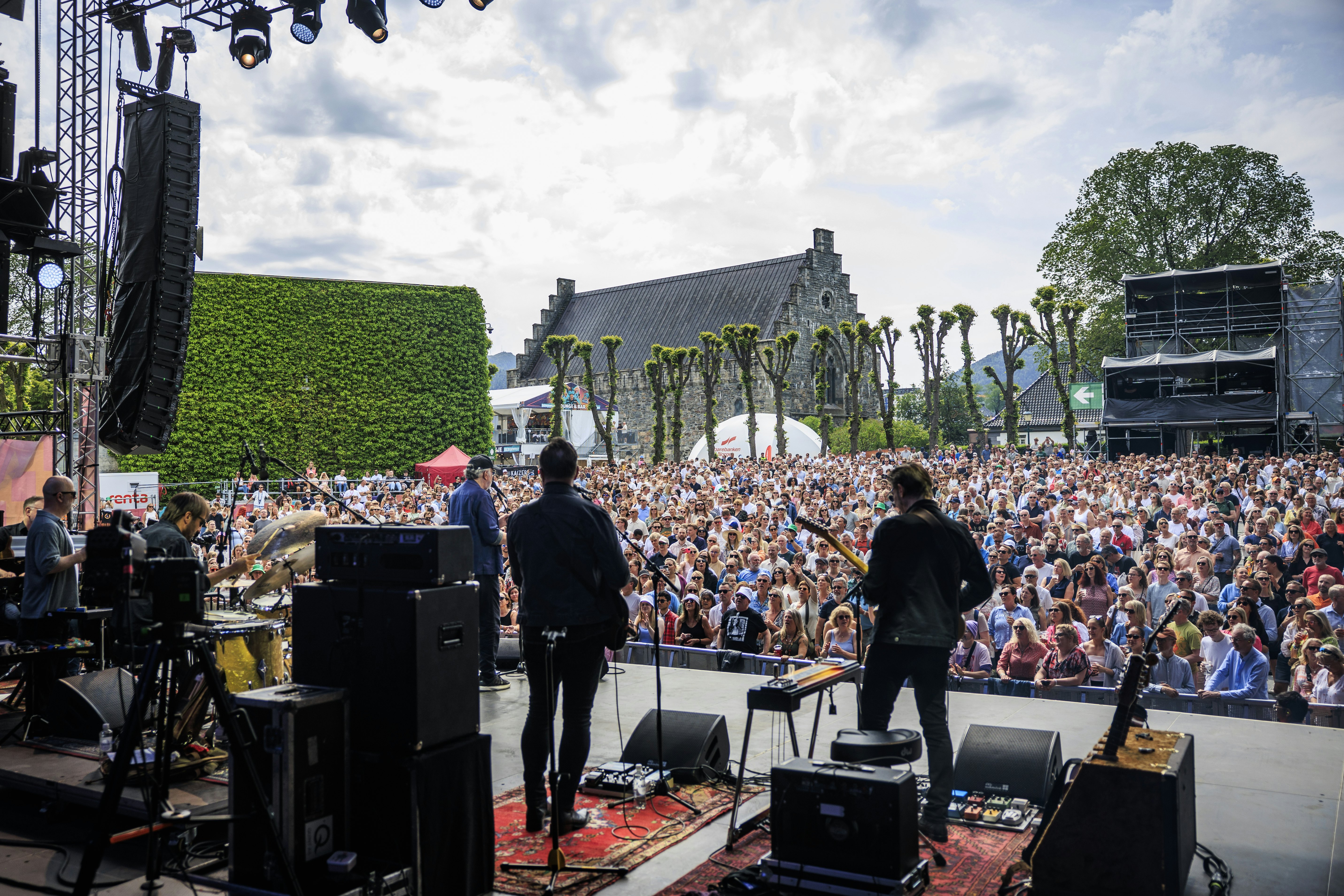 A band performs, pictured from behind, in front of a large crowd of spectators. Historic buildings are seen behind the crowd.