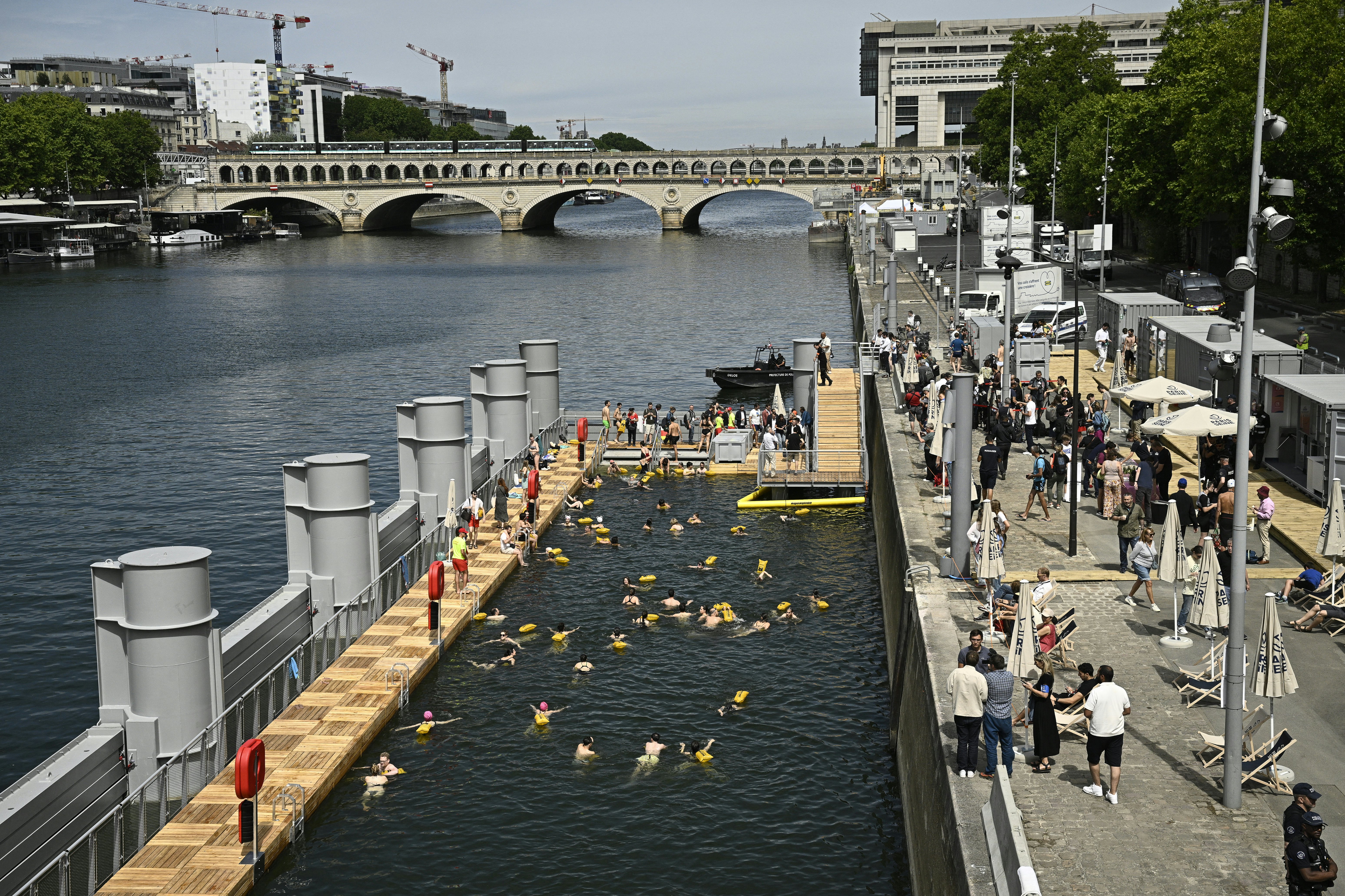 A wooden deck runs around the edge of a section of river to create a swimming pool within. All swimmers have a yellow floatation device attached to them, and lifeguards patrol the deck.