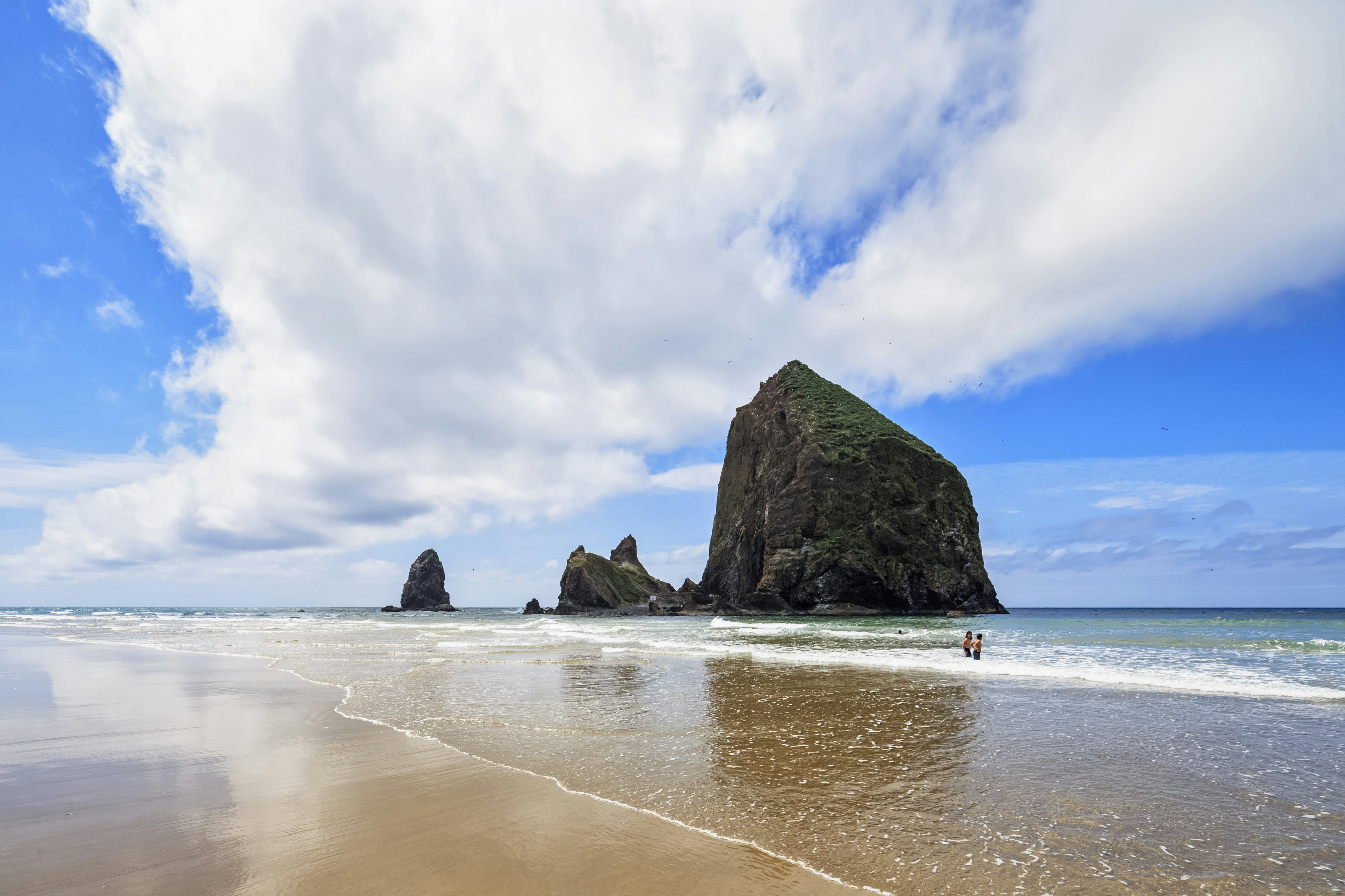 A large freestanding rock at the edge of the ocean. Two people paddle nearby.