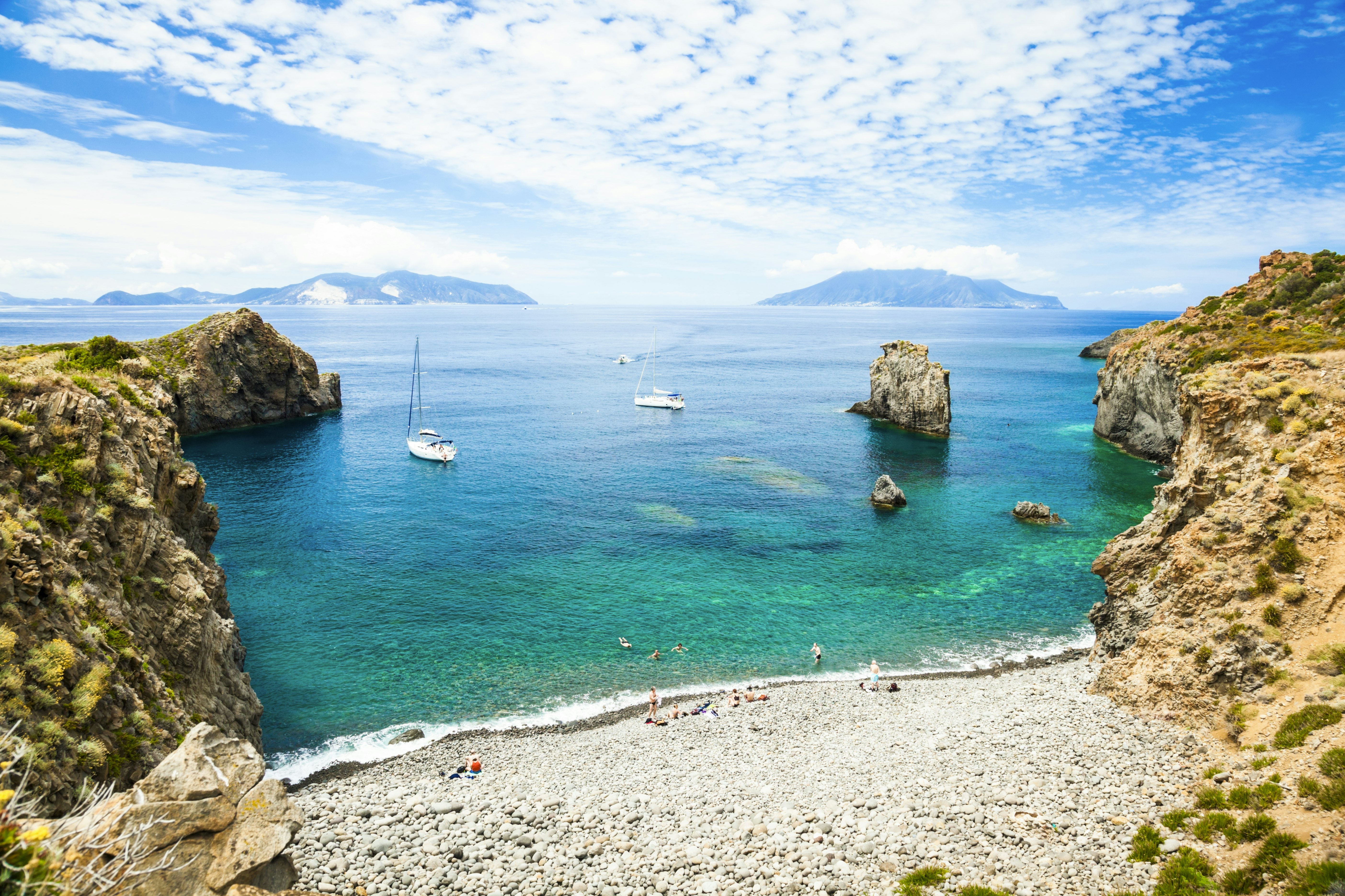Sail boats moored in the turquoise waters of a sheltered bay with a pebbly beach. People swim in the sea and sunbathe.