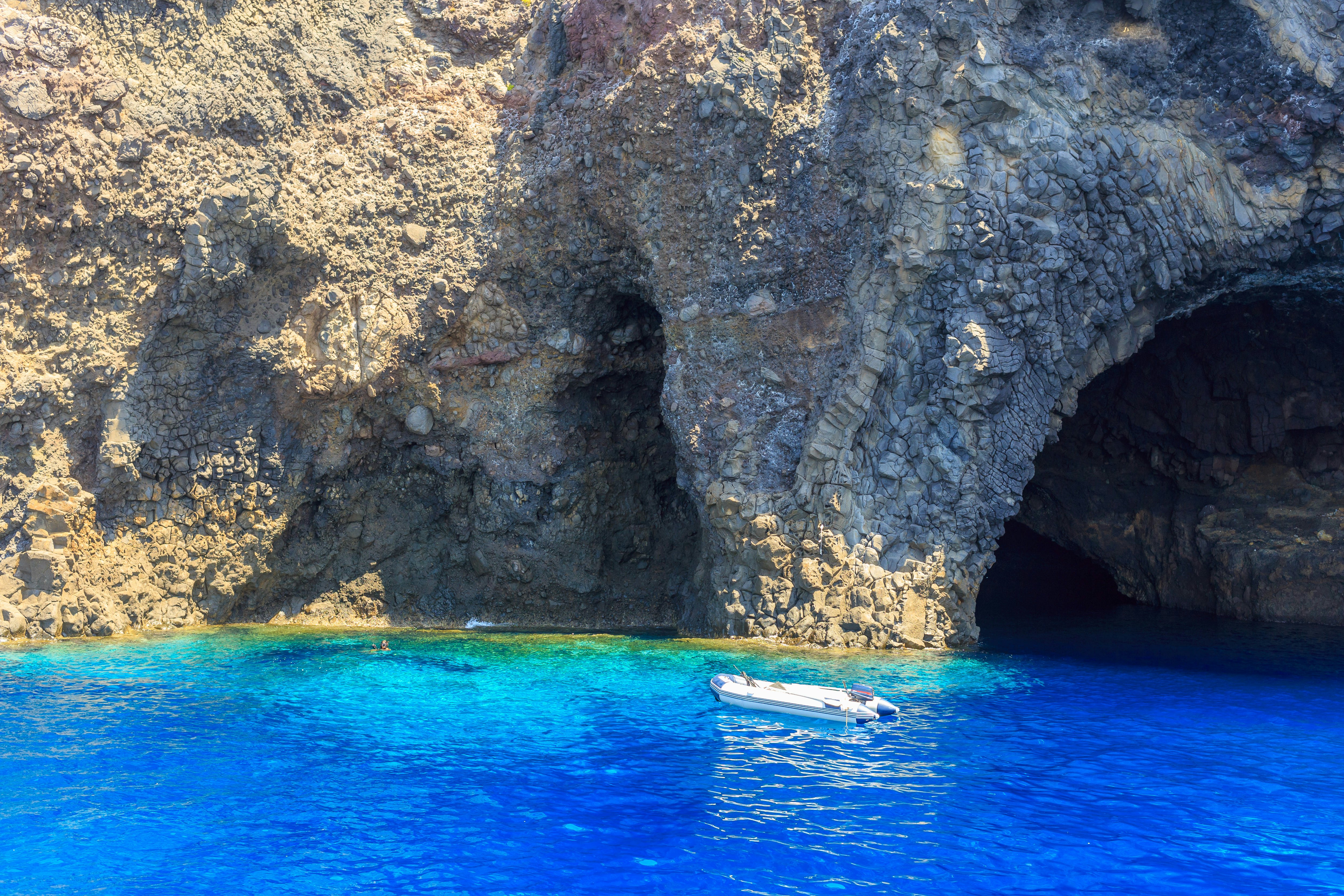 A small motorized boat moored in blue ocean near a cave formation. Two swimmers take a dip near the rocky coastline.