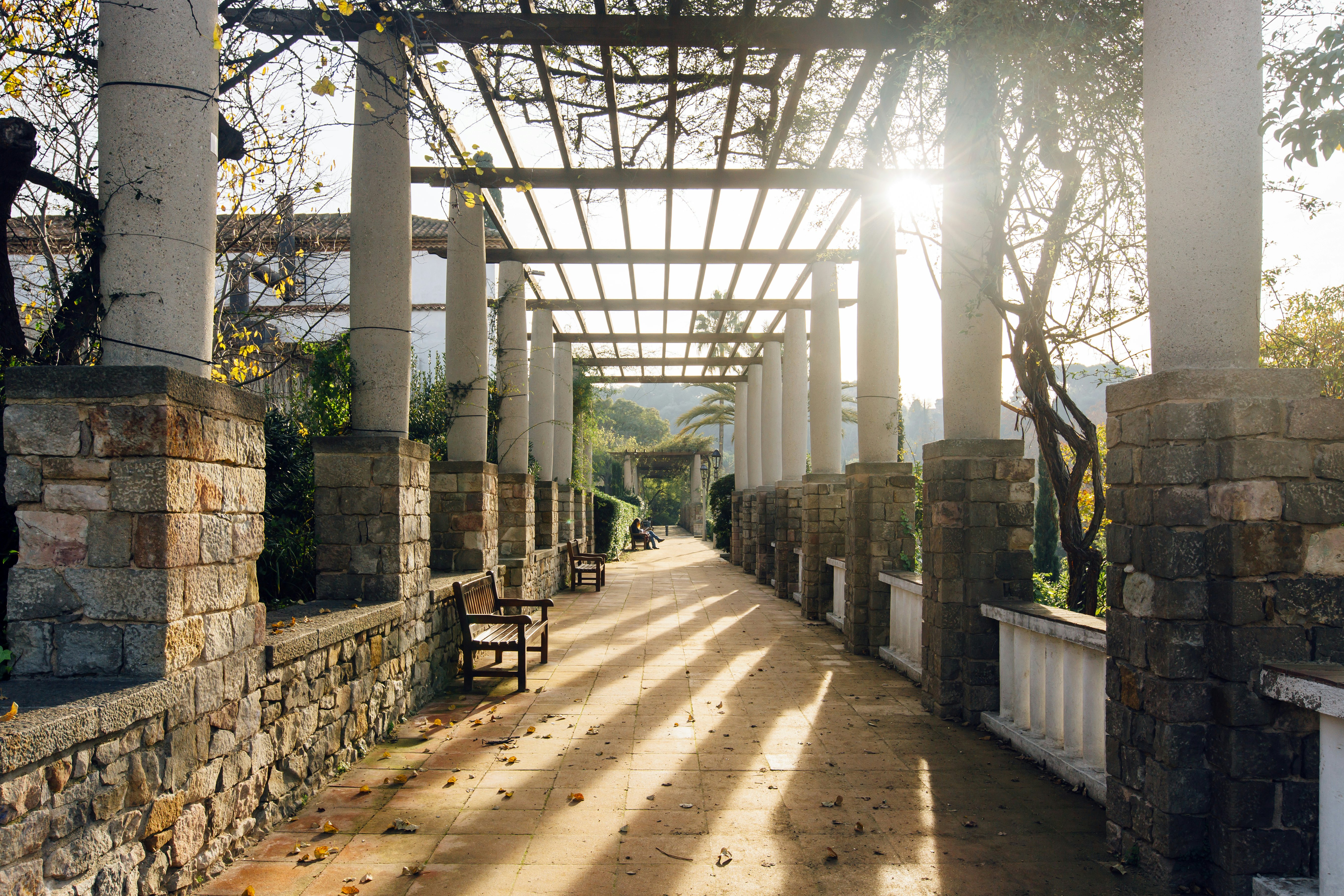 Sun shining through the slats of a portico in a park in Barcelona; there are several empty benches with people sitting in the distance.