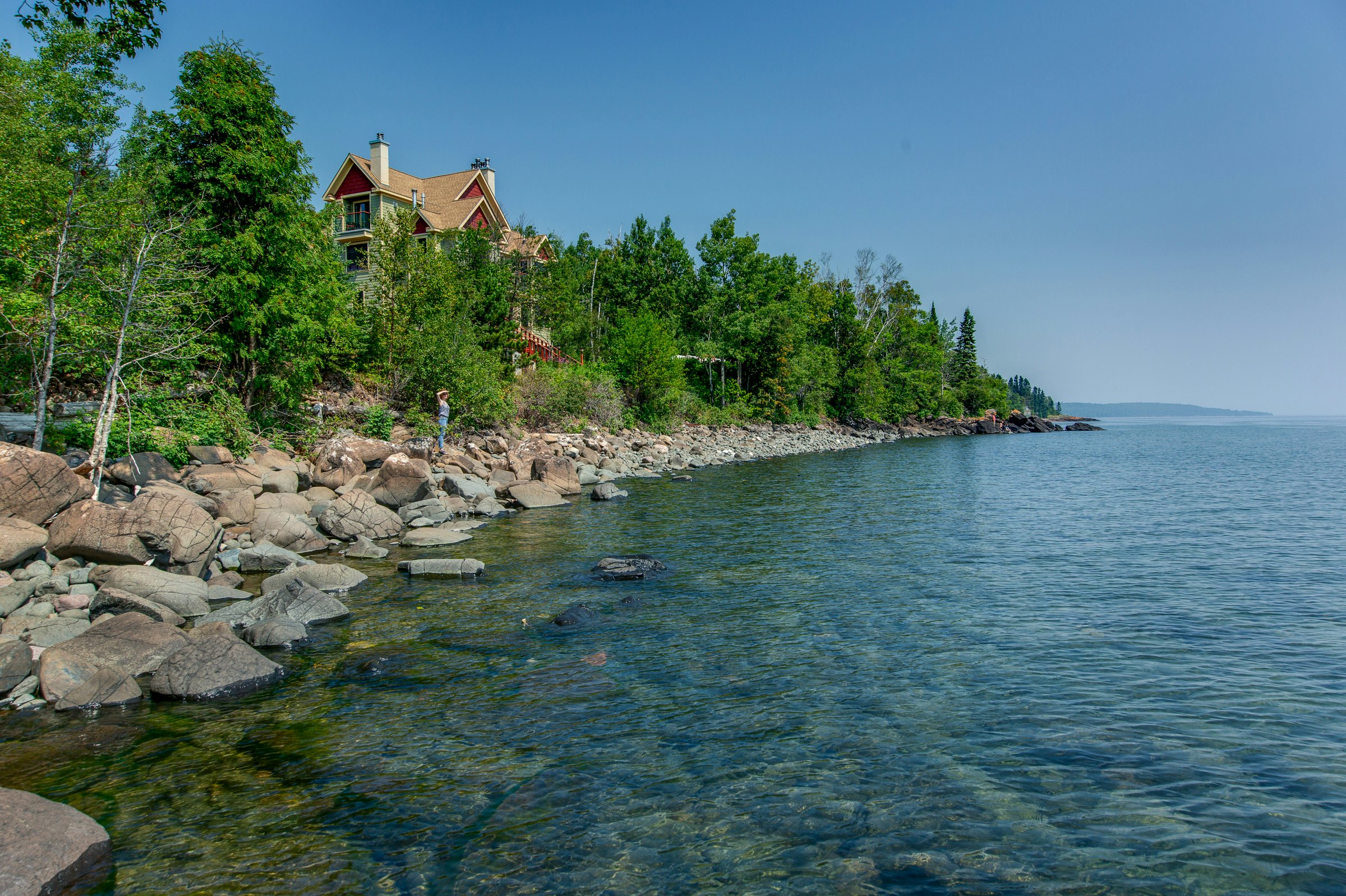 Blue rocky water with a person on shore and tall trees in front of a house in the background on Lake Superior, Minnesota