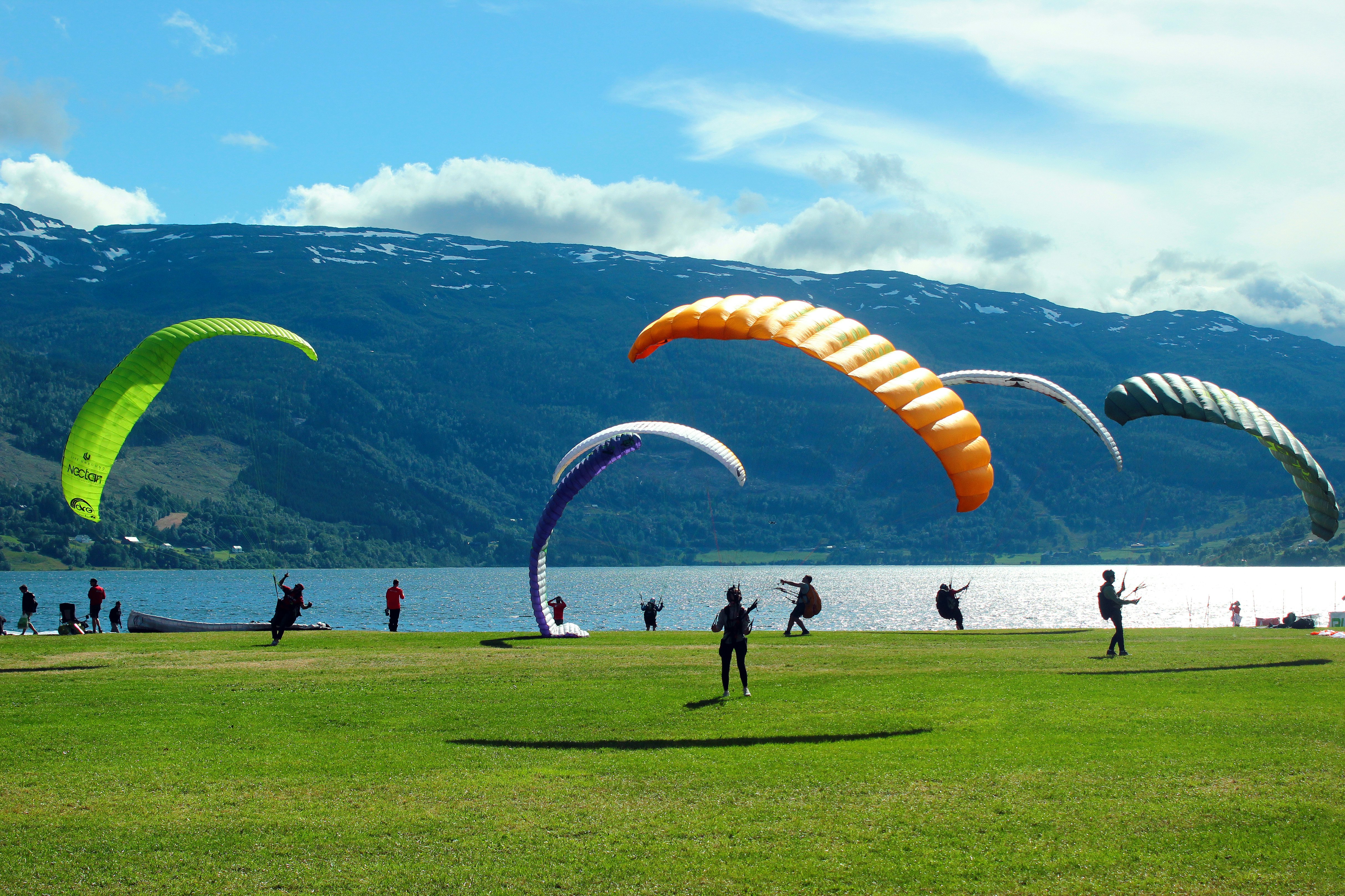Paragliders with parachutes in bright colors stand on a lawn in front of a lake, with mountains in the distance.