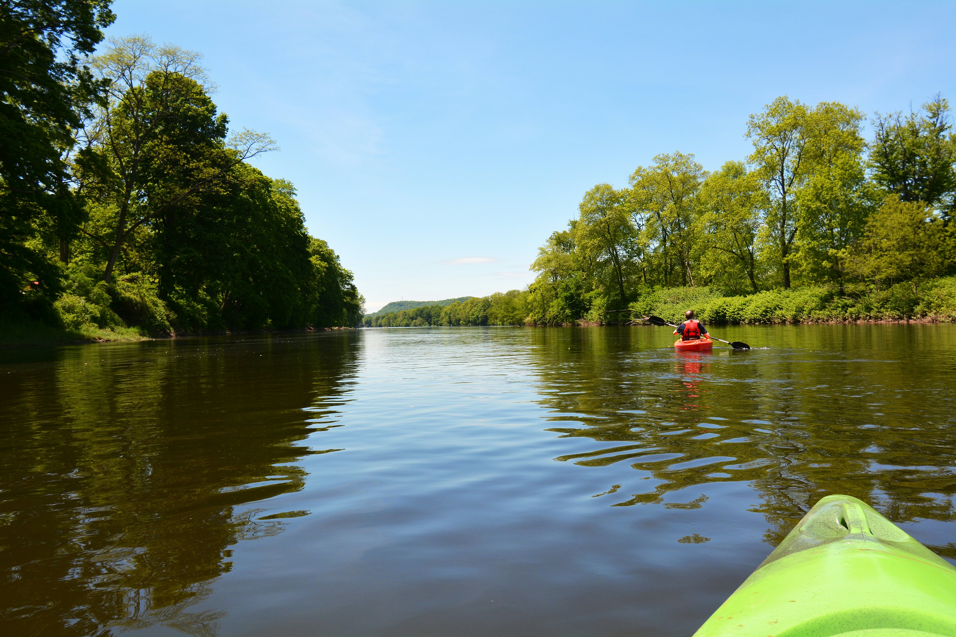 The tip of a yellow-green kayak in the foreground, with a person in a red life vest and red kayak in the middle-ground on the Delaware River