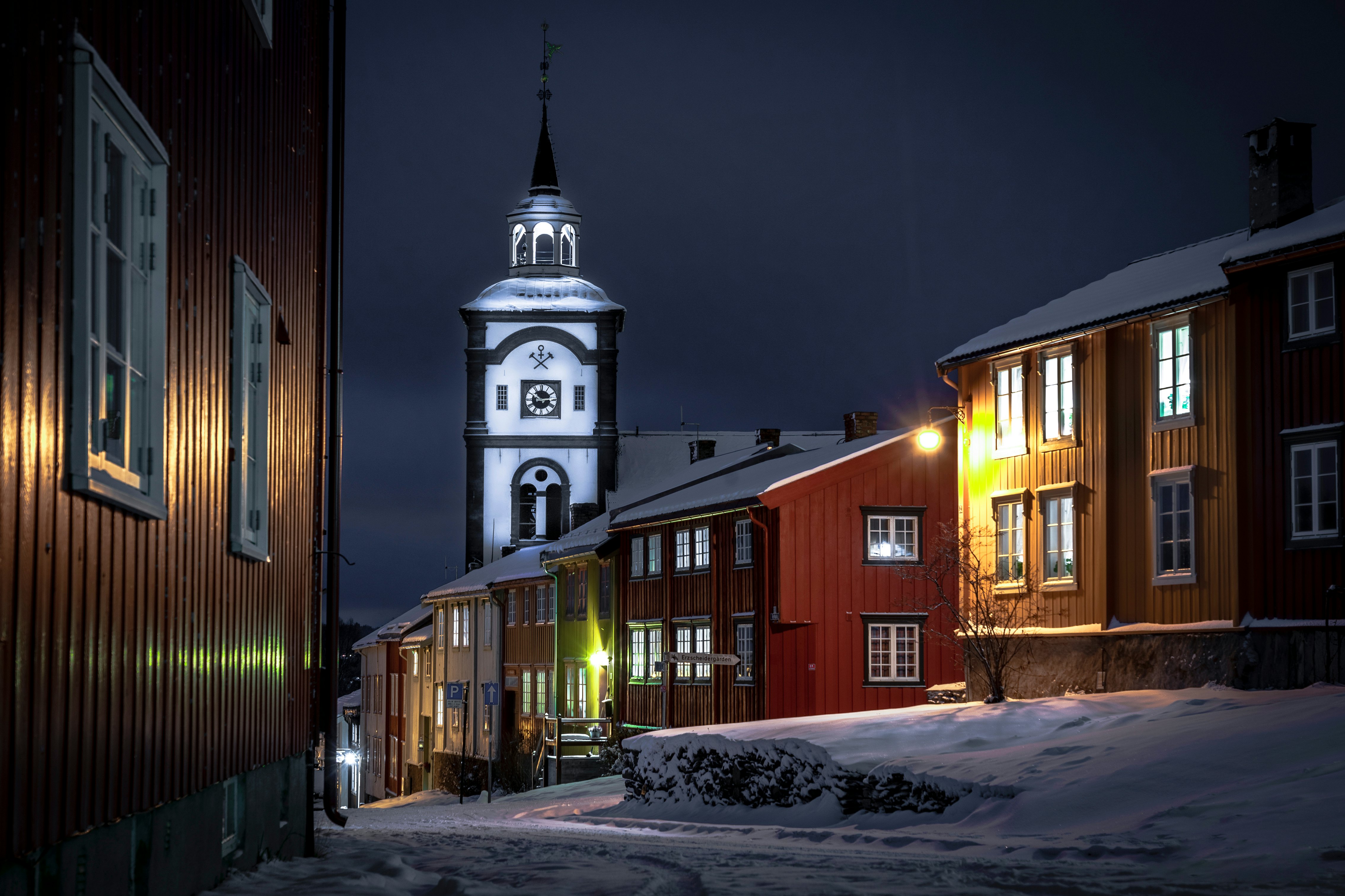 The white tower of a church is seen at night in a historic village. Snow blankets the ground.