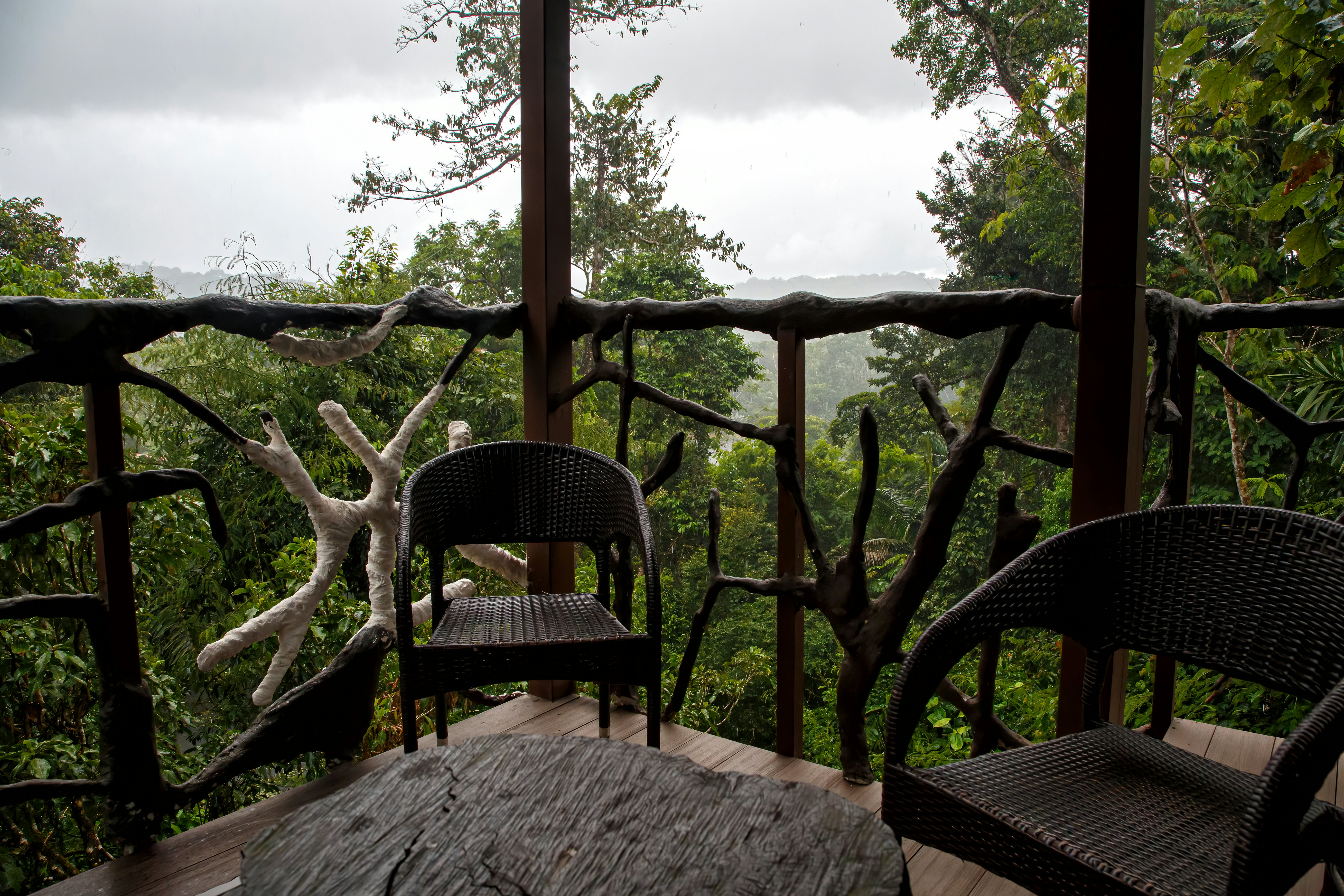 Two dark woven chairs and a woodblock table are on a deck with carved wooden railings that's above the trees in Costa Rica.
