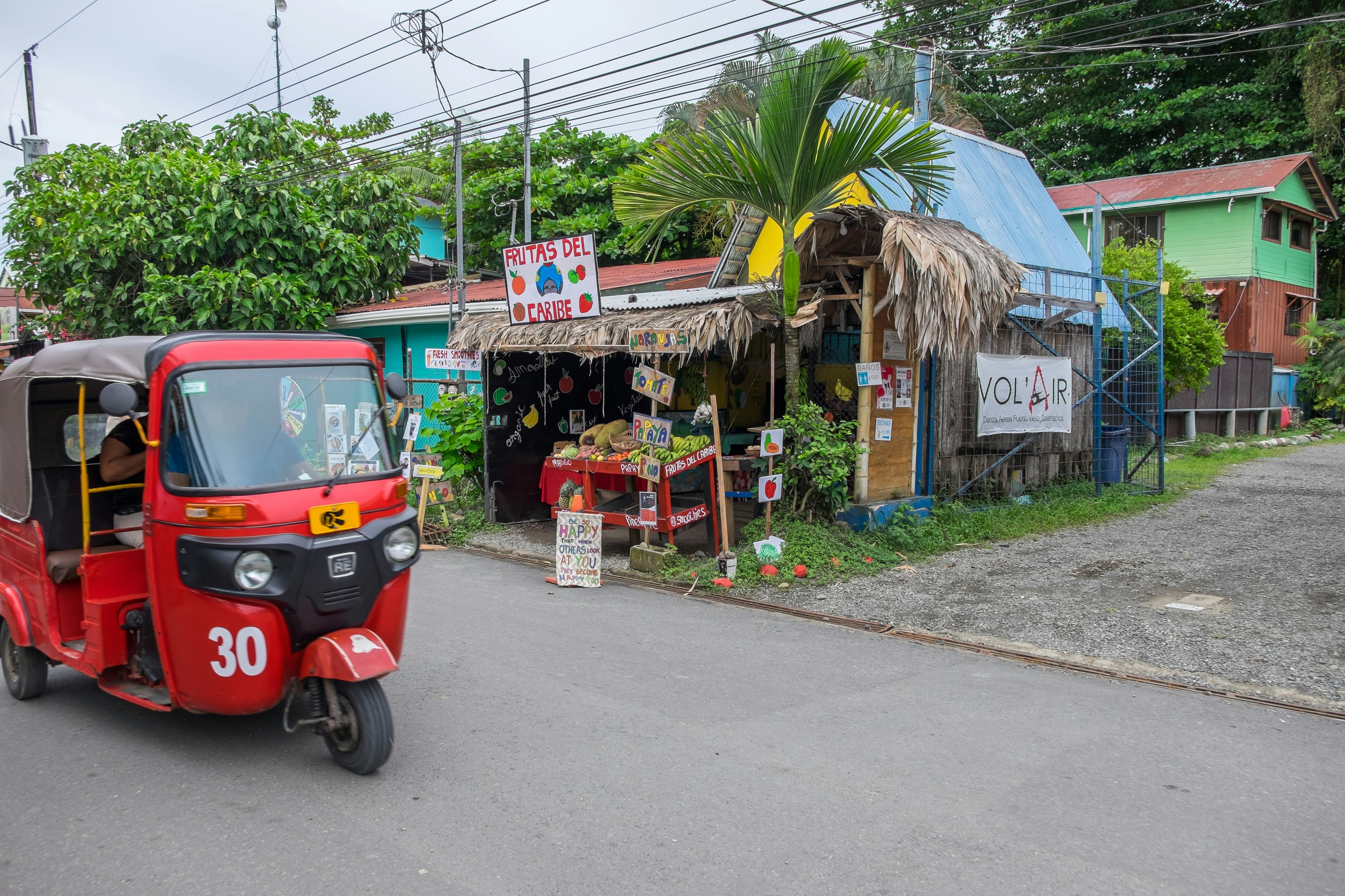 A red three-wheeled vehicle drives on a road past a fruit stand in Costa Rica.