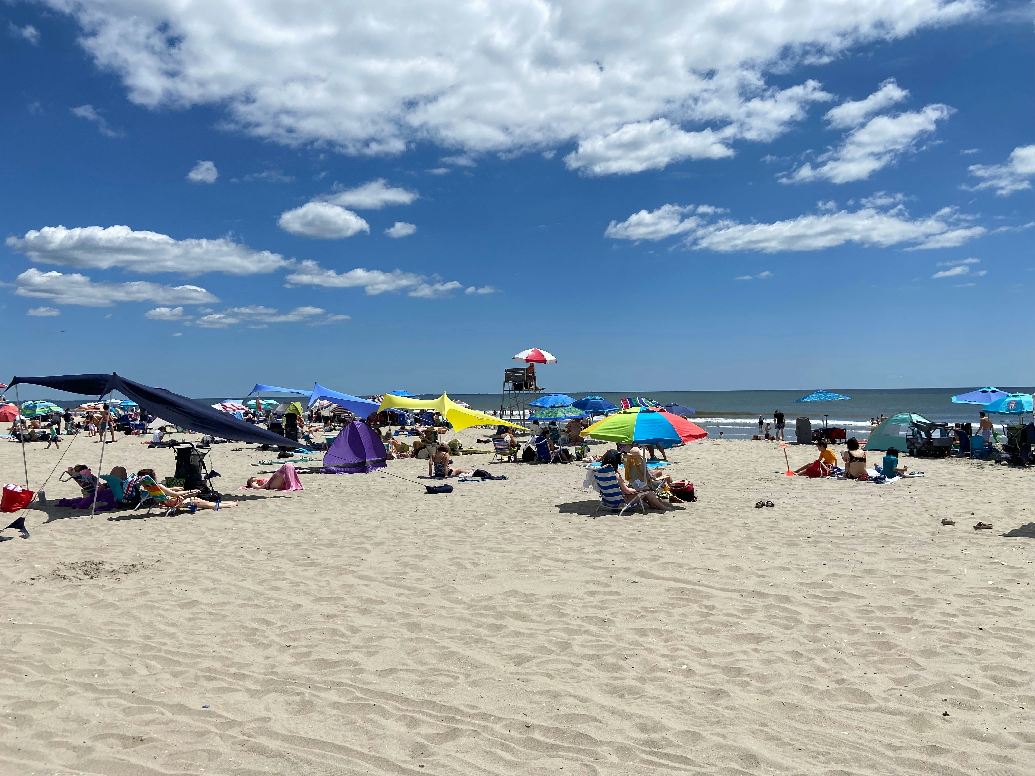 A crowded beach with a lifeguard under an umbrella surrounded by colorful tents and sun shades on a sunny summer day.