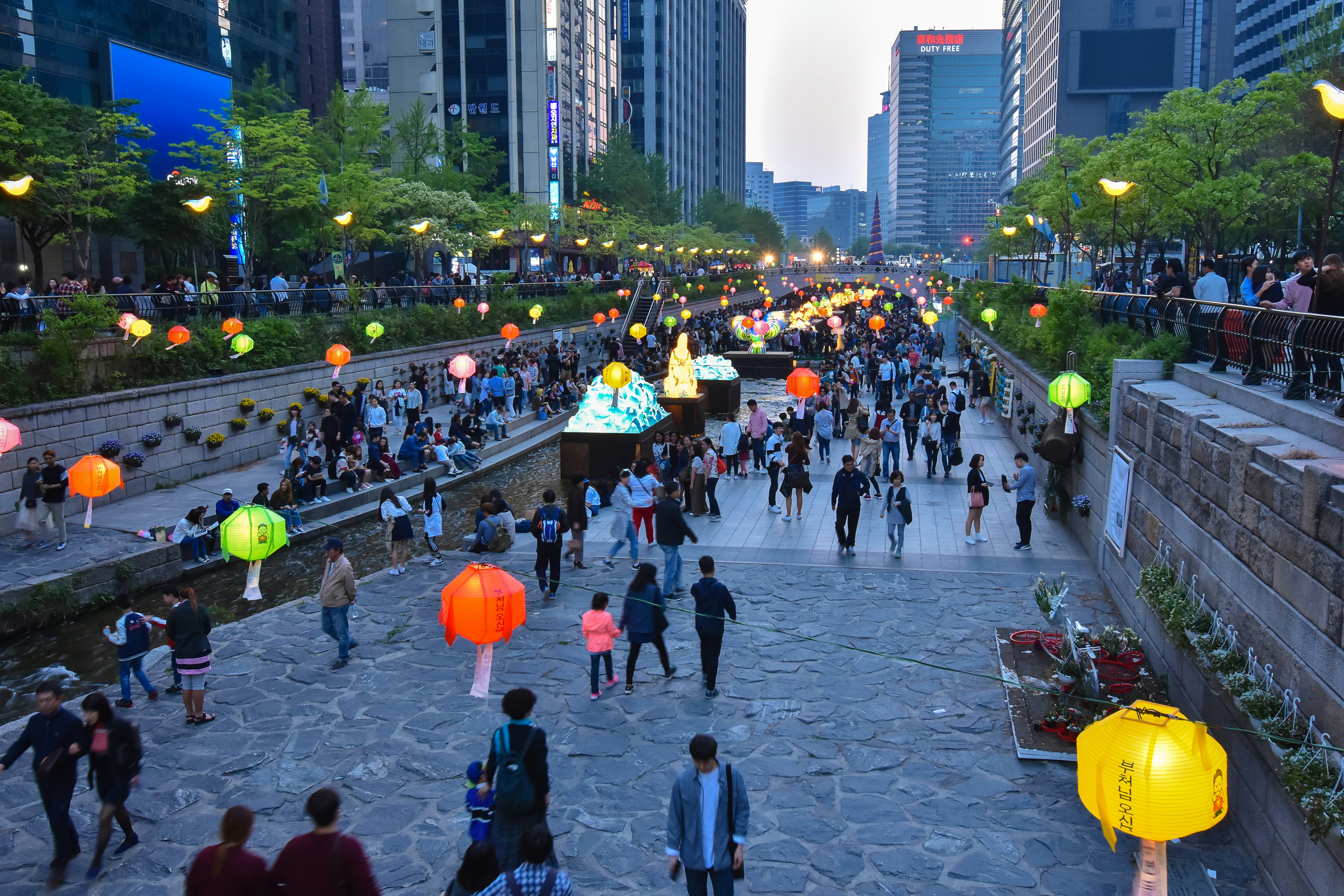 Paper lanterns hang over a public park and promenade in the built-up central business district of a city.