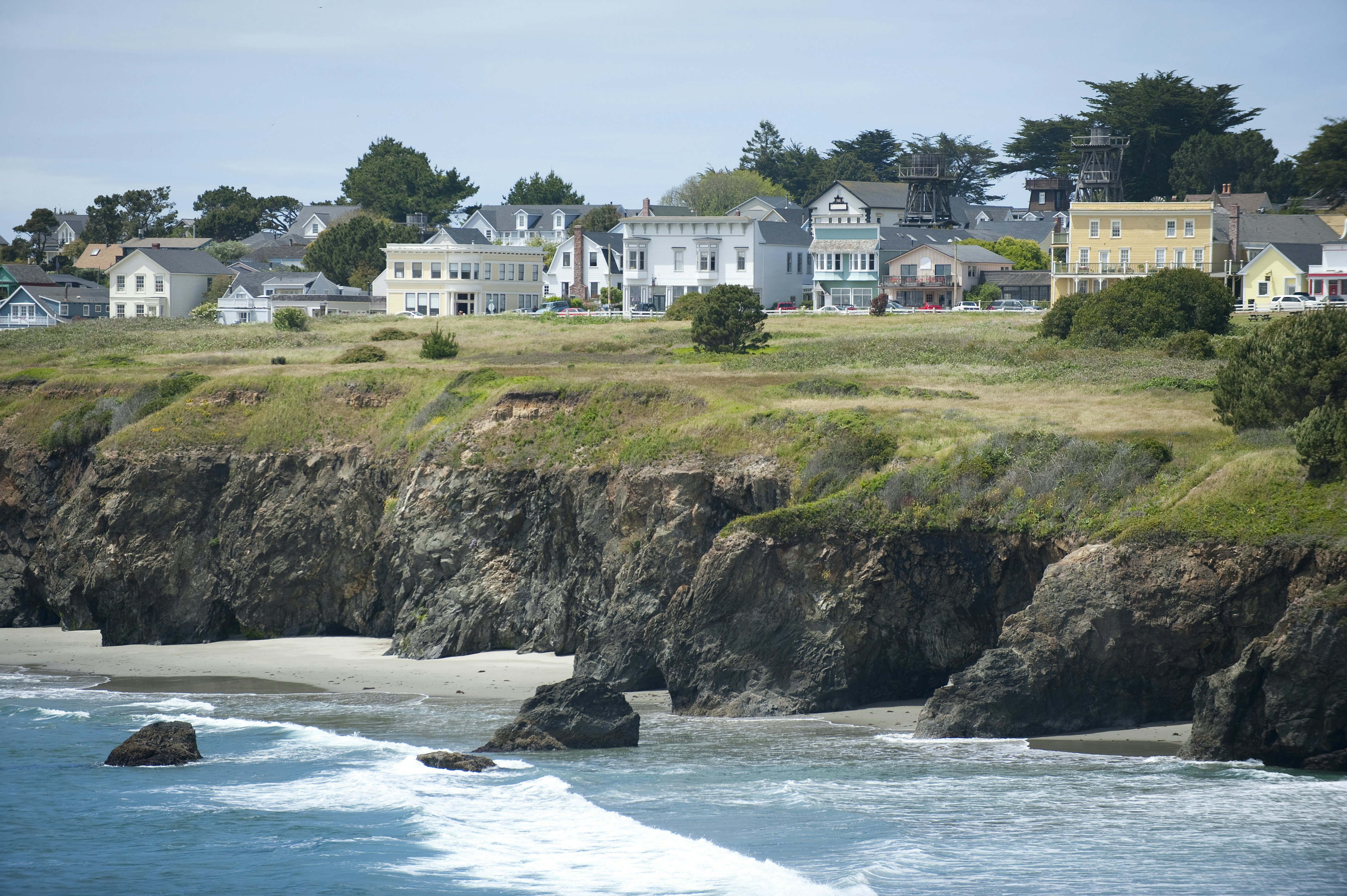 Low buildings are set back from a grassy cliff by the ocean in California.