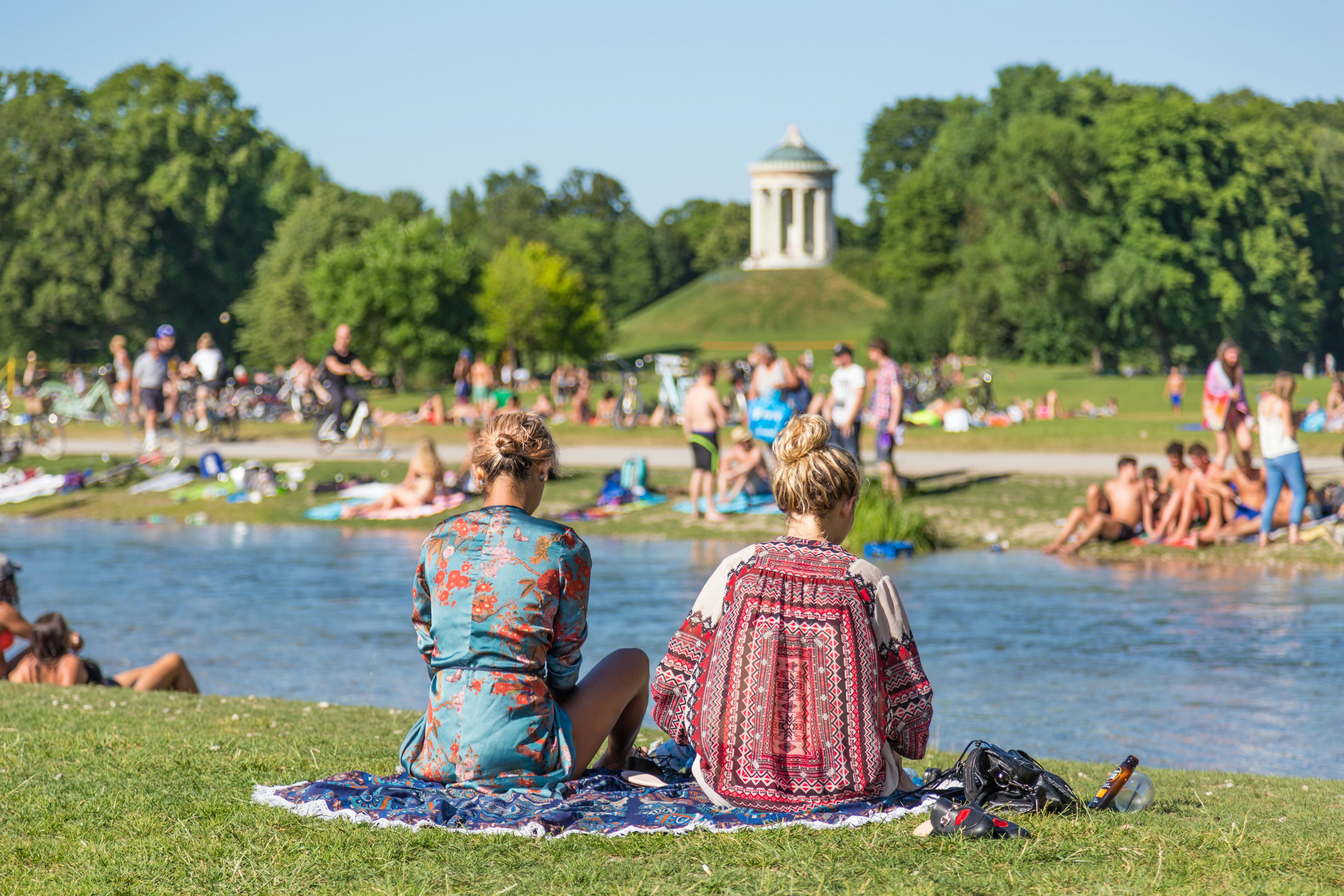 Two women sitting on a blanket in a park are seen from behind in front of a pond. Other people surrounding them lounge and sunbathe; a temple-like structure is seen on a knoll in the distance.