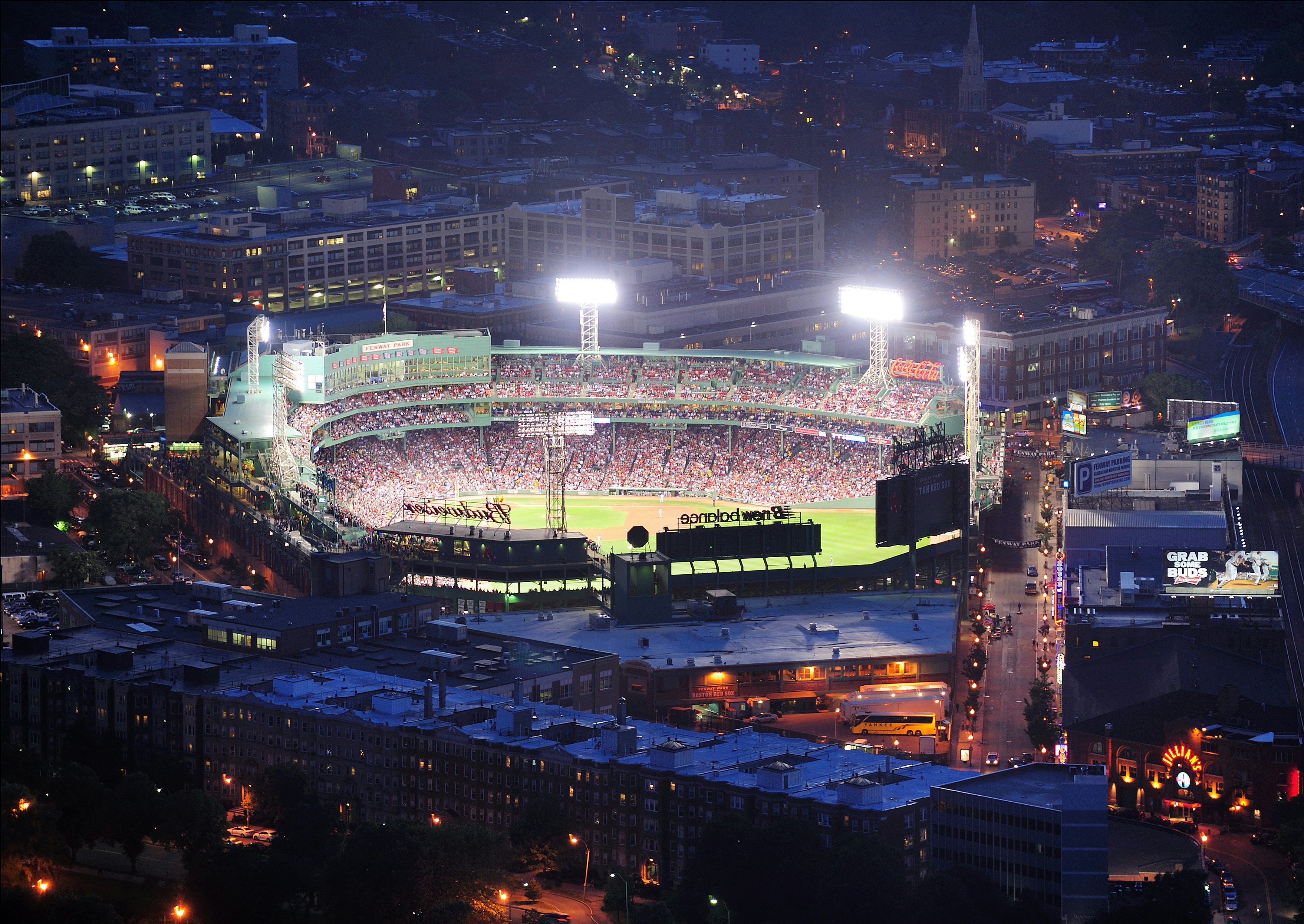 An aerial view of a baseball stadium in a city, with a night game taking place.
