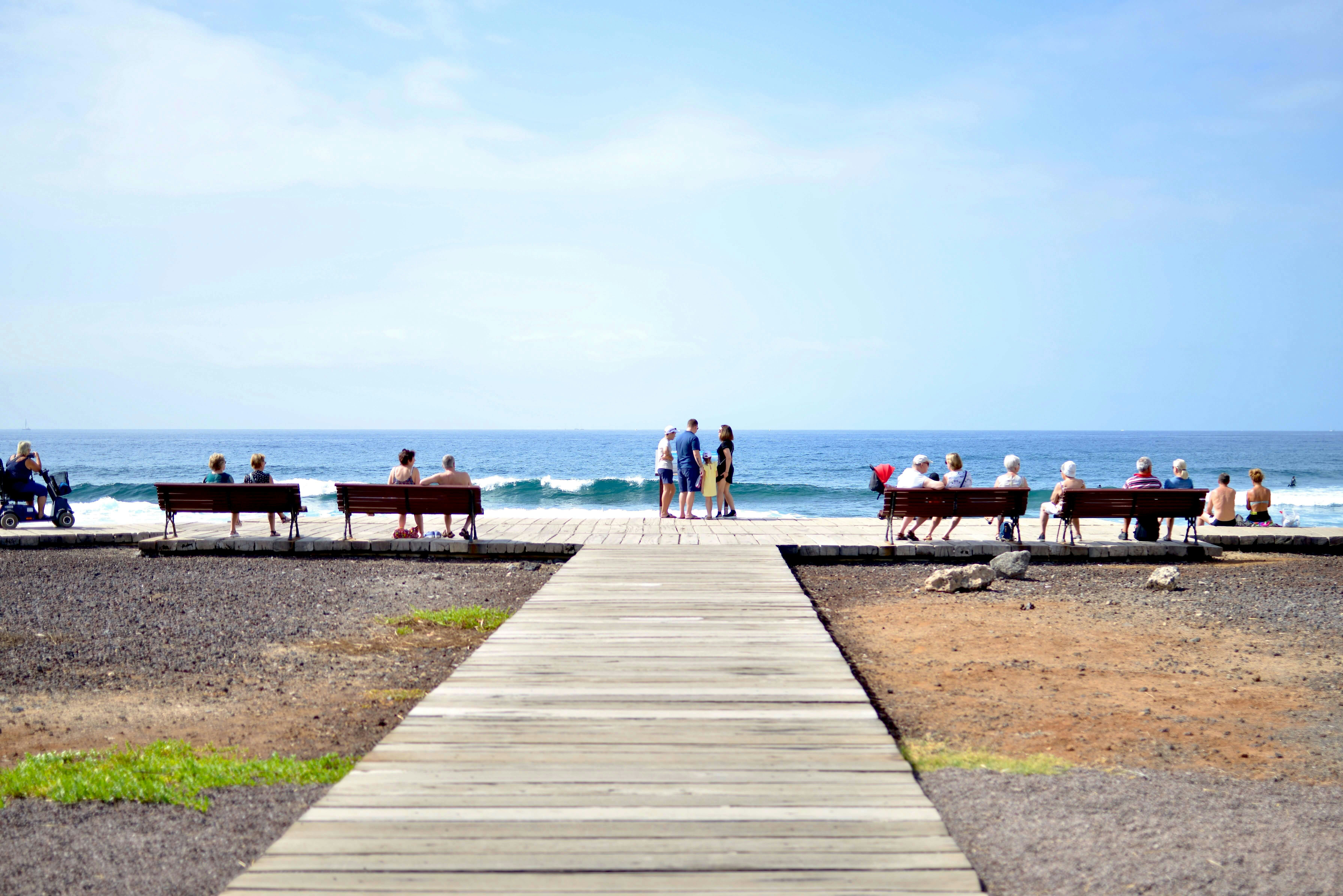 People relaxing on benches on a promenade in front of waves on a beach with blue sky and sea