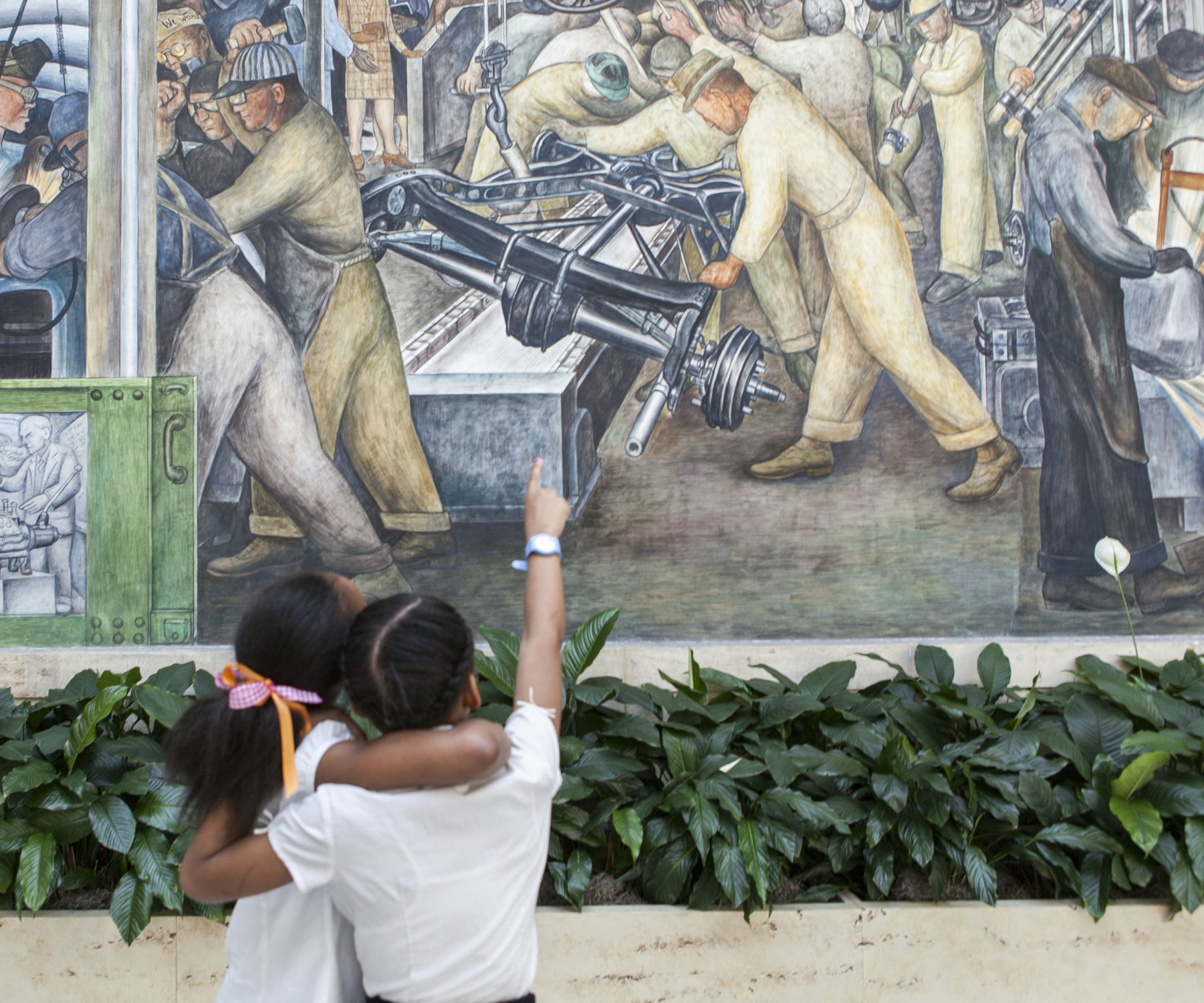 Two young children with arms around each other and one pointing up at a large mural by Diego Rivera with green peace lily flowers in front inside a museum.