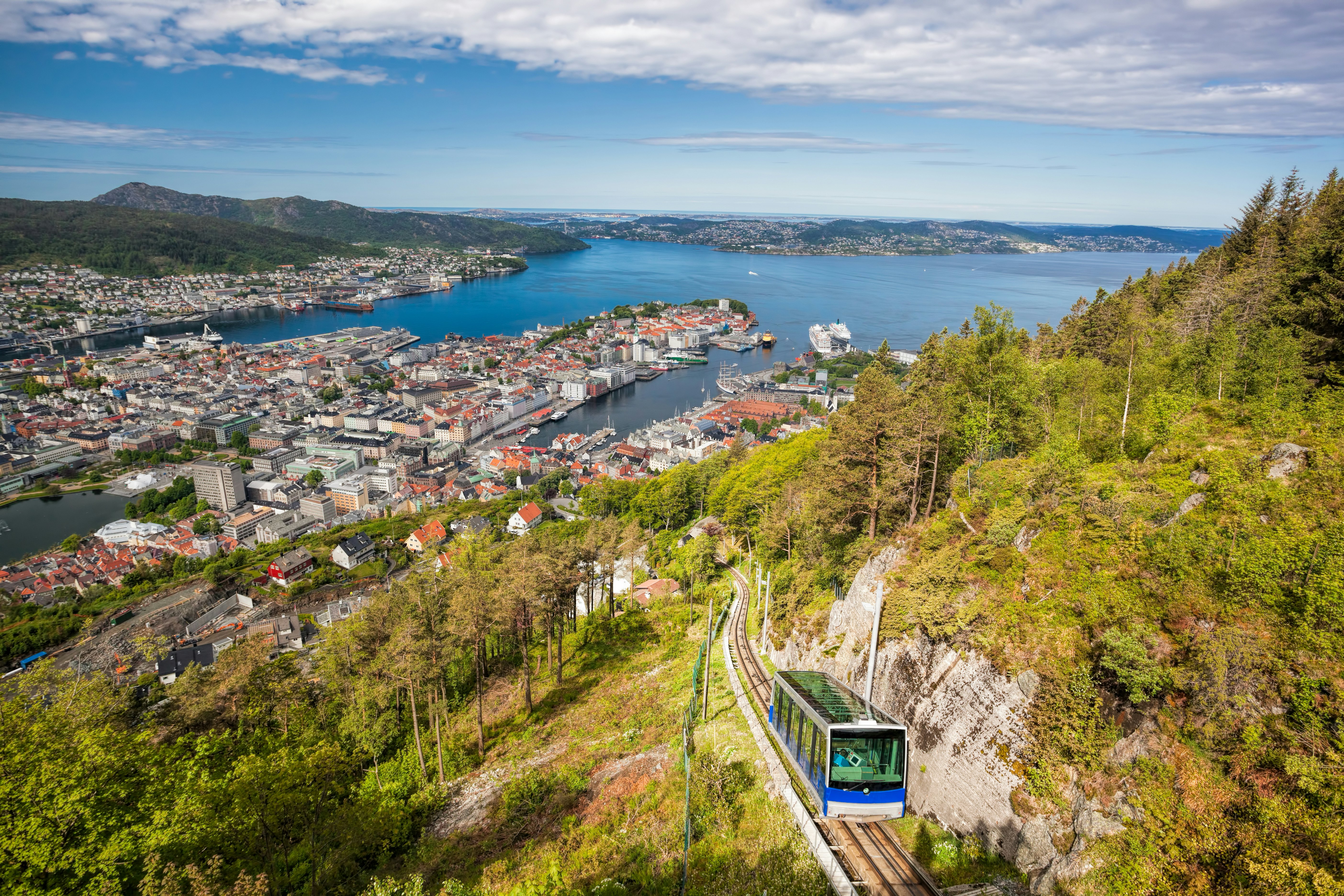 A blue funicular car ascends a tree-covered hill as a city is visible below in the distance.