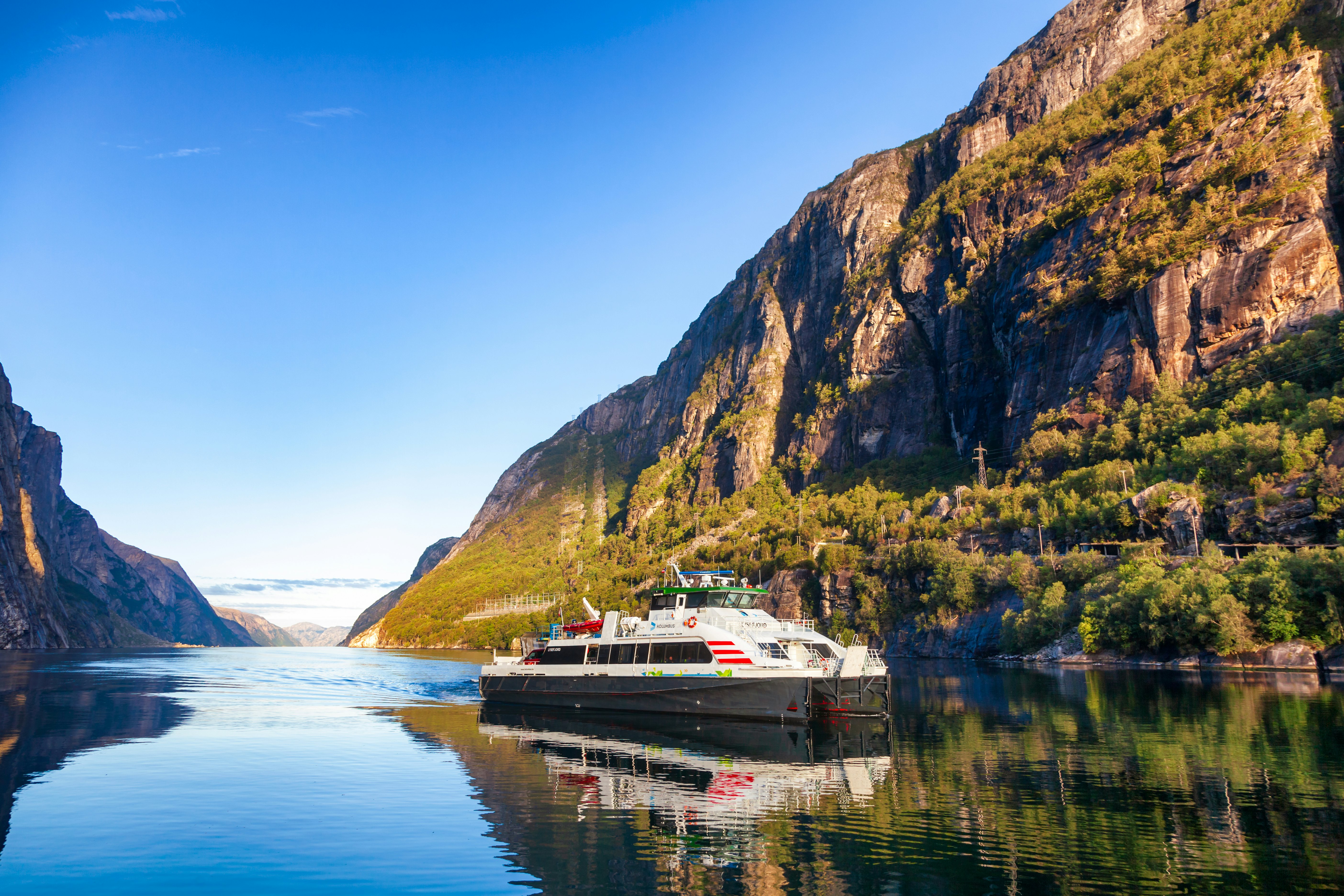 A boat plies the calm waters of a fjord. Tree-covered cliffs rise on either side of the water.