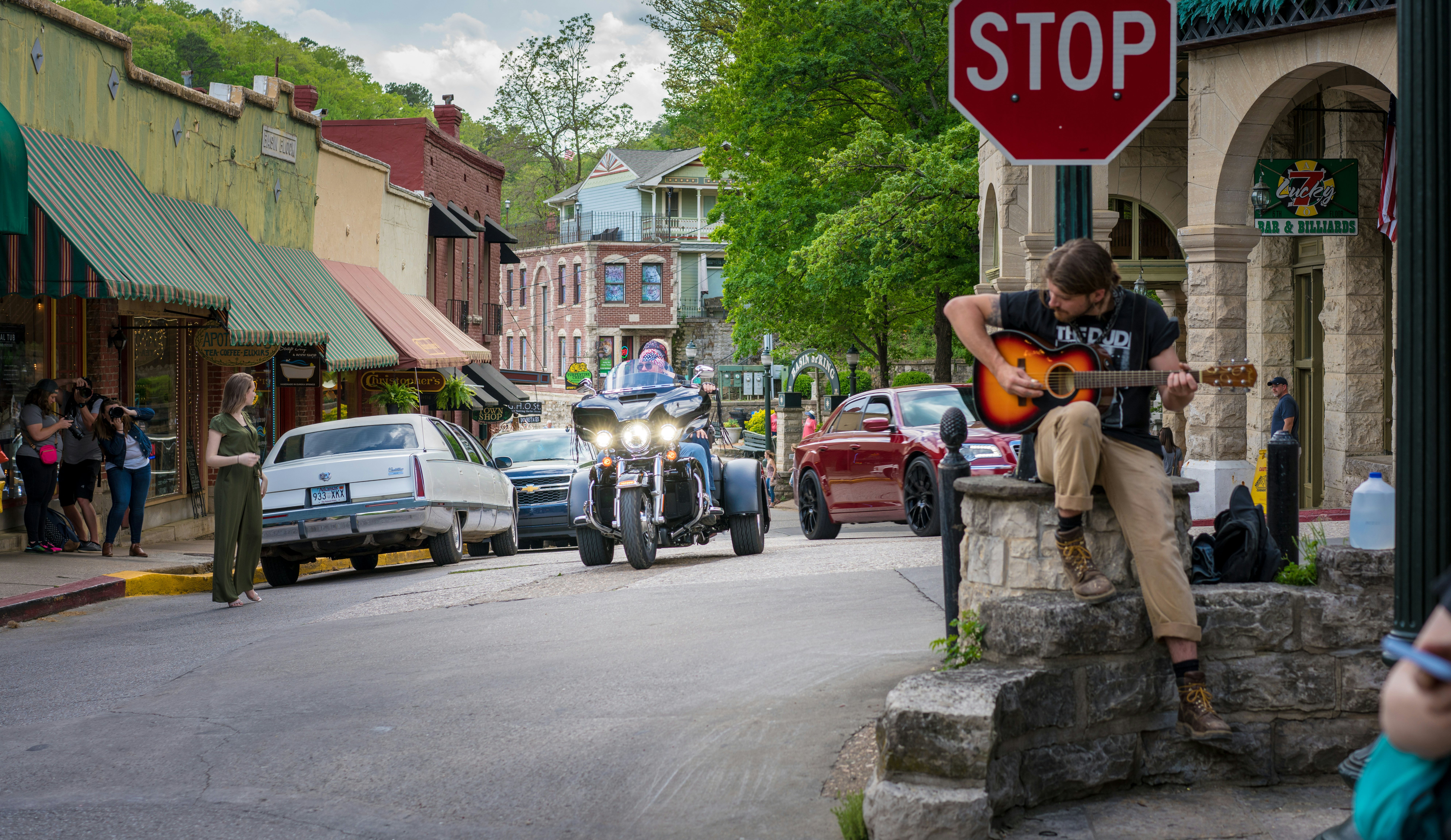 A biker in a three-wheel vehicle drives down the street of a town. A musician with a guitar is seen on a porch to the right.