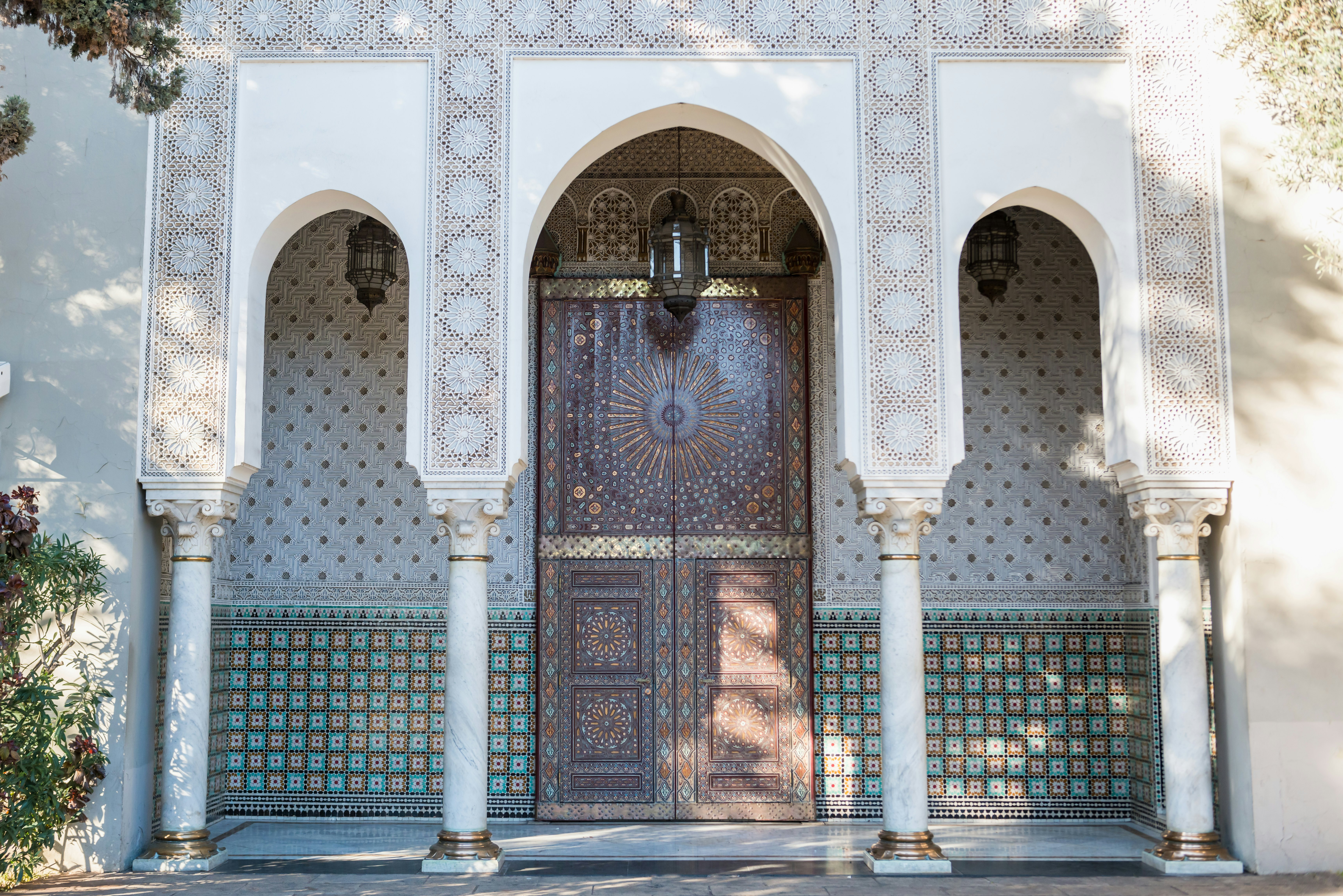 An elaborately decorated wood door set back in an alcove covered in patterned tiles is reached through an archway with four carved columns.