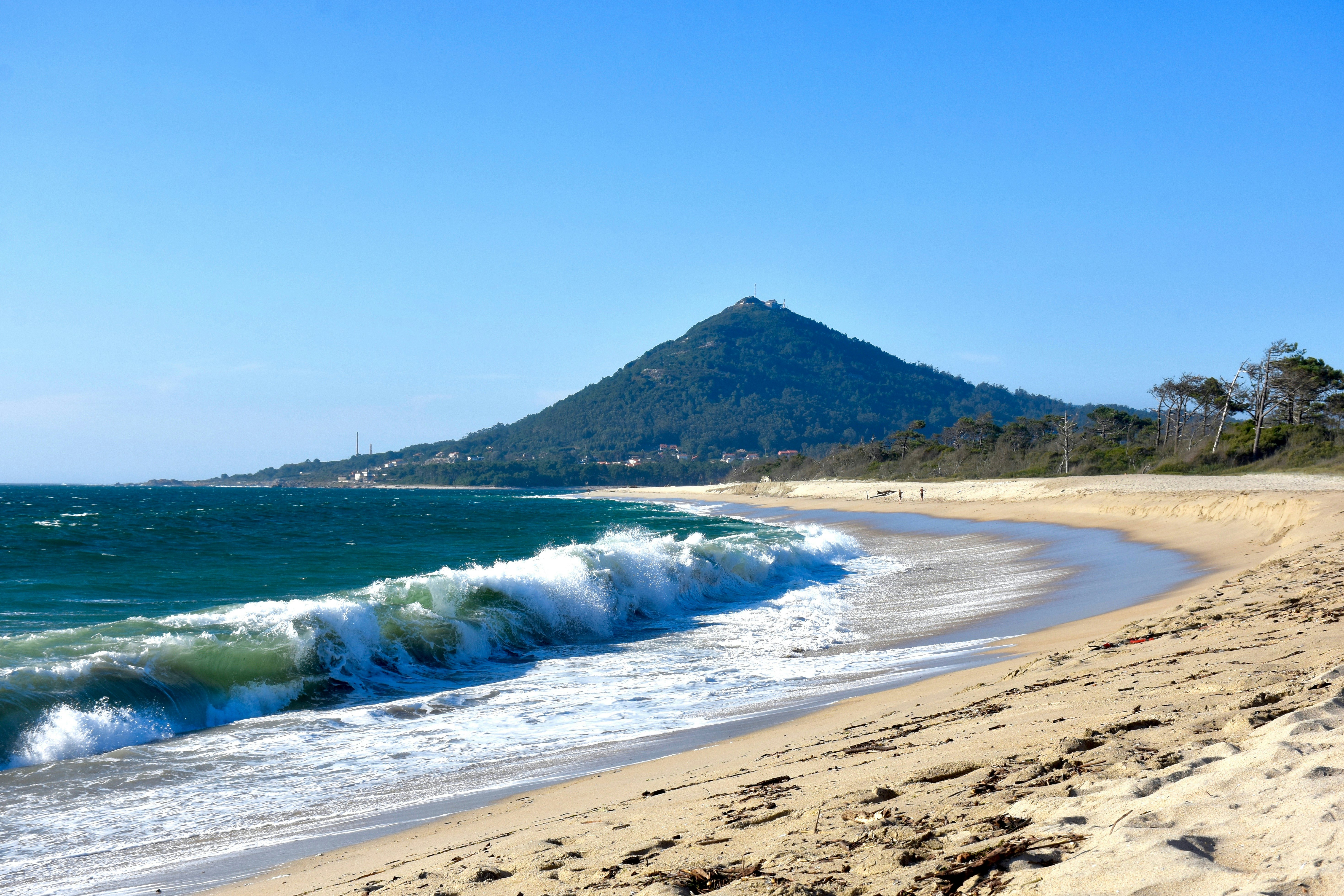 White-capped waves rolling onto a curved sandy beach with a conical forested mountain in the distance on a sunny day.