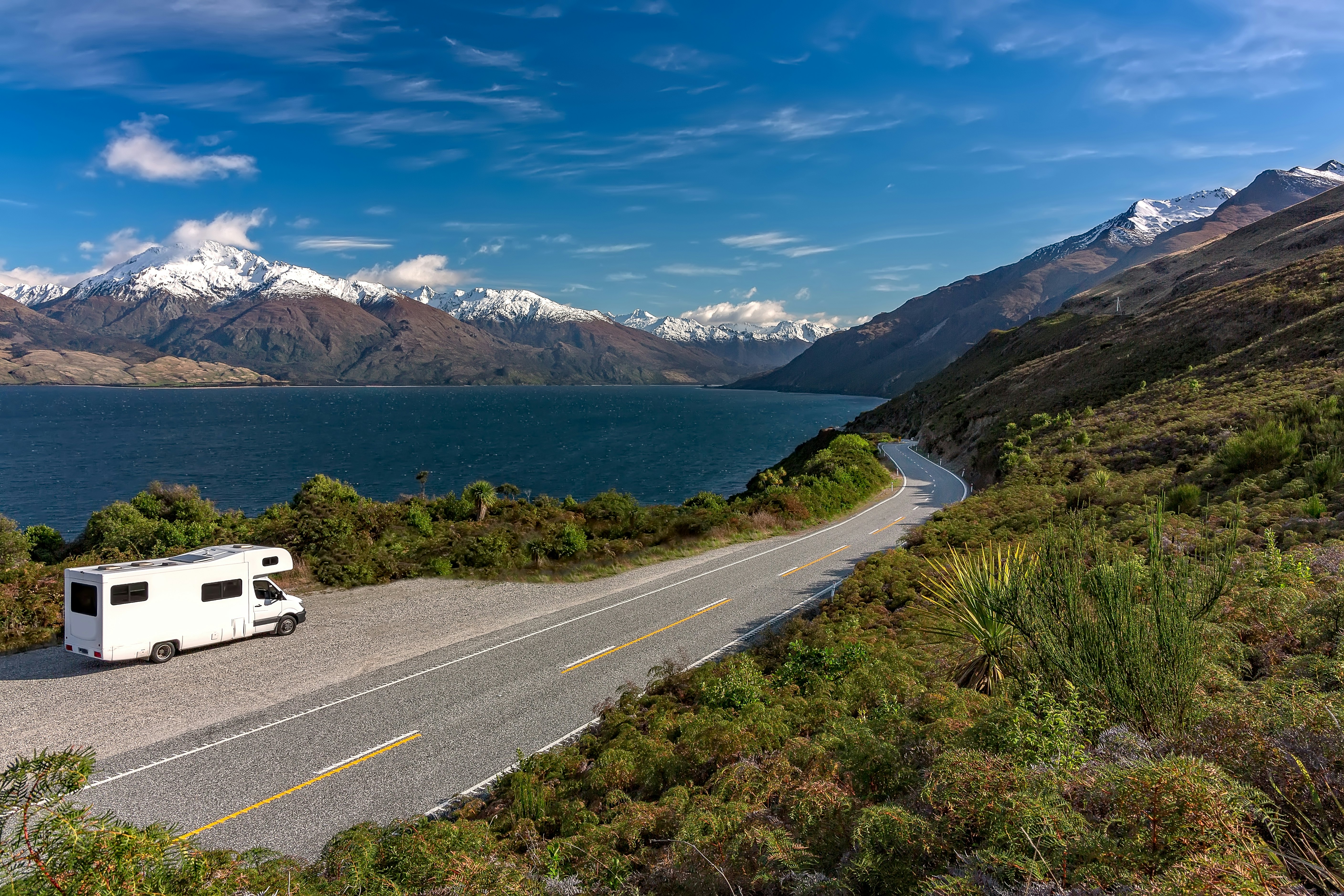 A white camper-van parked in a lay-by viewpoint on the banks of Lake Wānaka with the lake and the dramatic snow capped peaks in the distance, New Zealand.