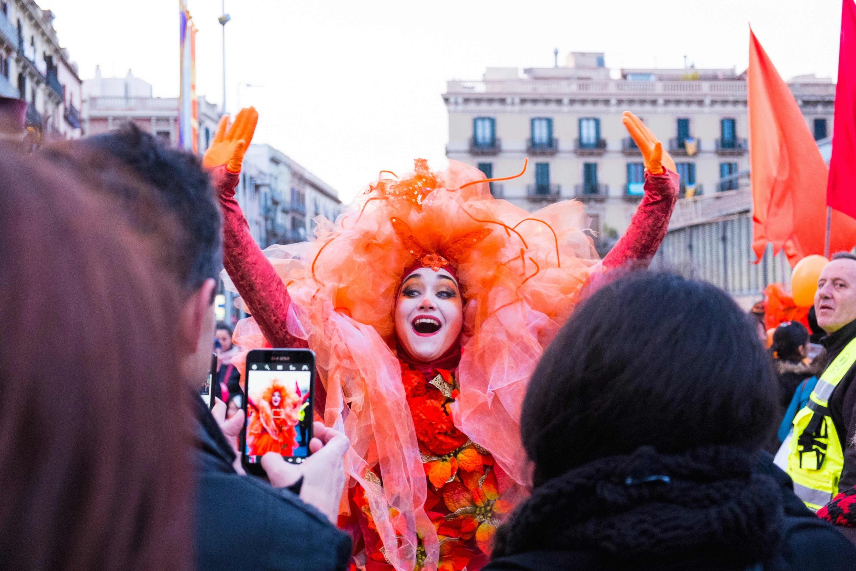 A performer in the Carnival dressed all in shades of orange and with her face painted poses for a photograph with her hands in the air.