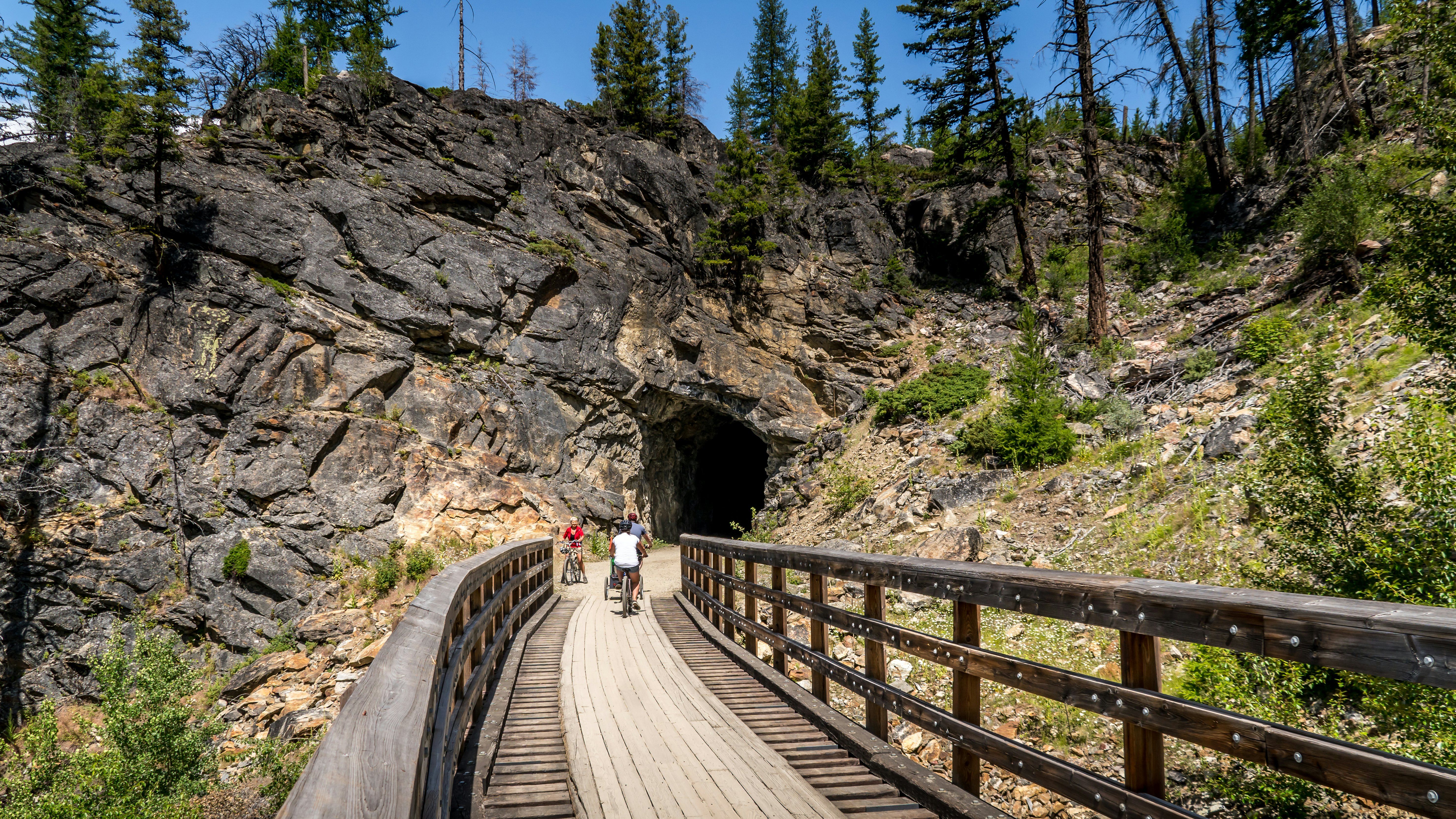 A cyclist pedals over wooden trestle bridge heading towards a tunnel through rock on a former rail line.