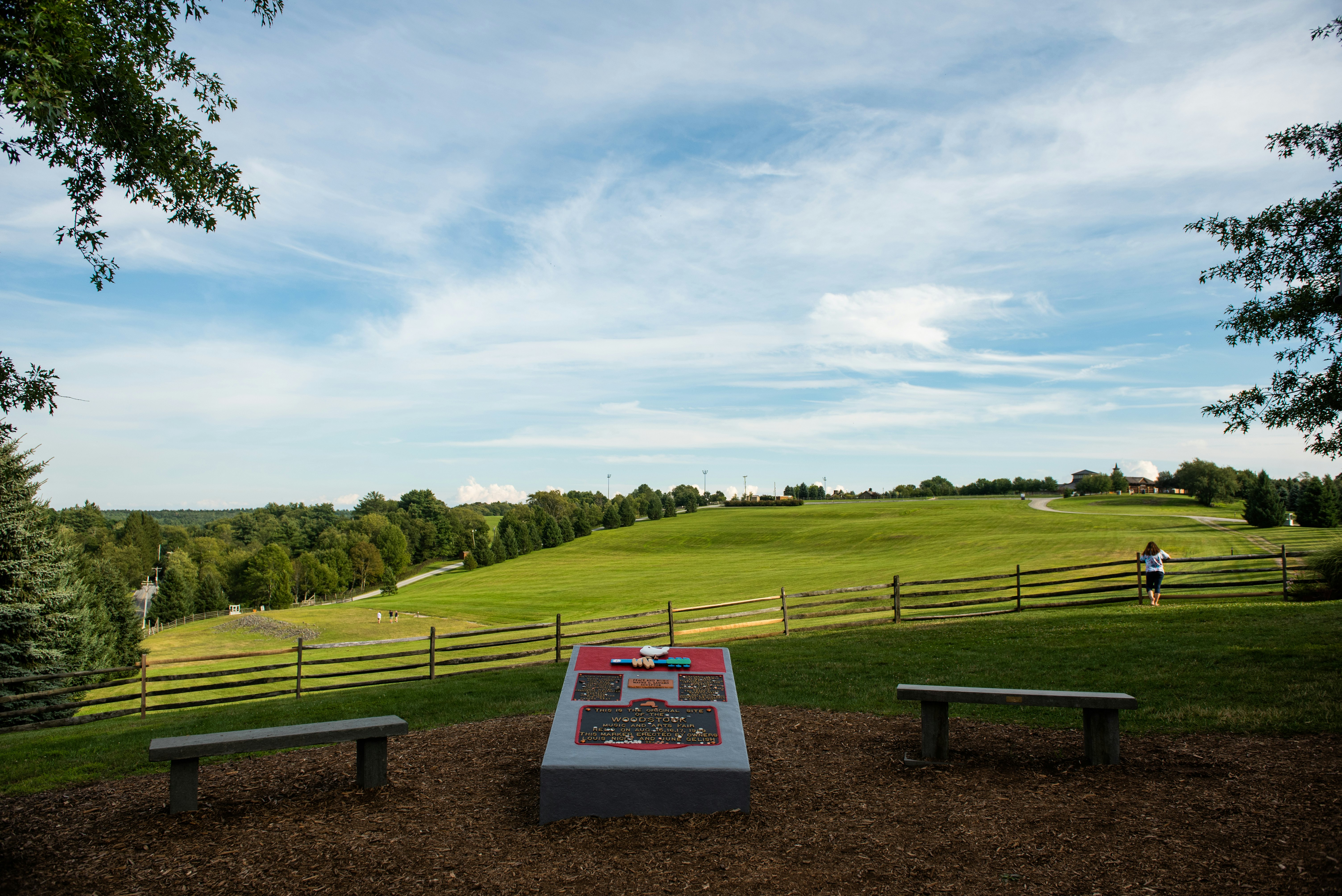 Two benches flank a large stone monument, with a fence and rolling green hills in the background