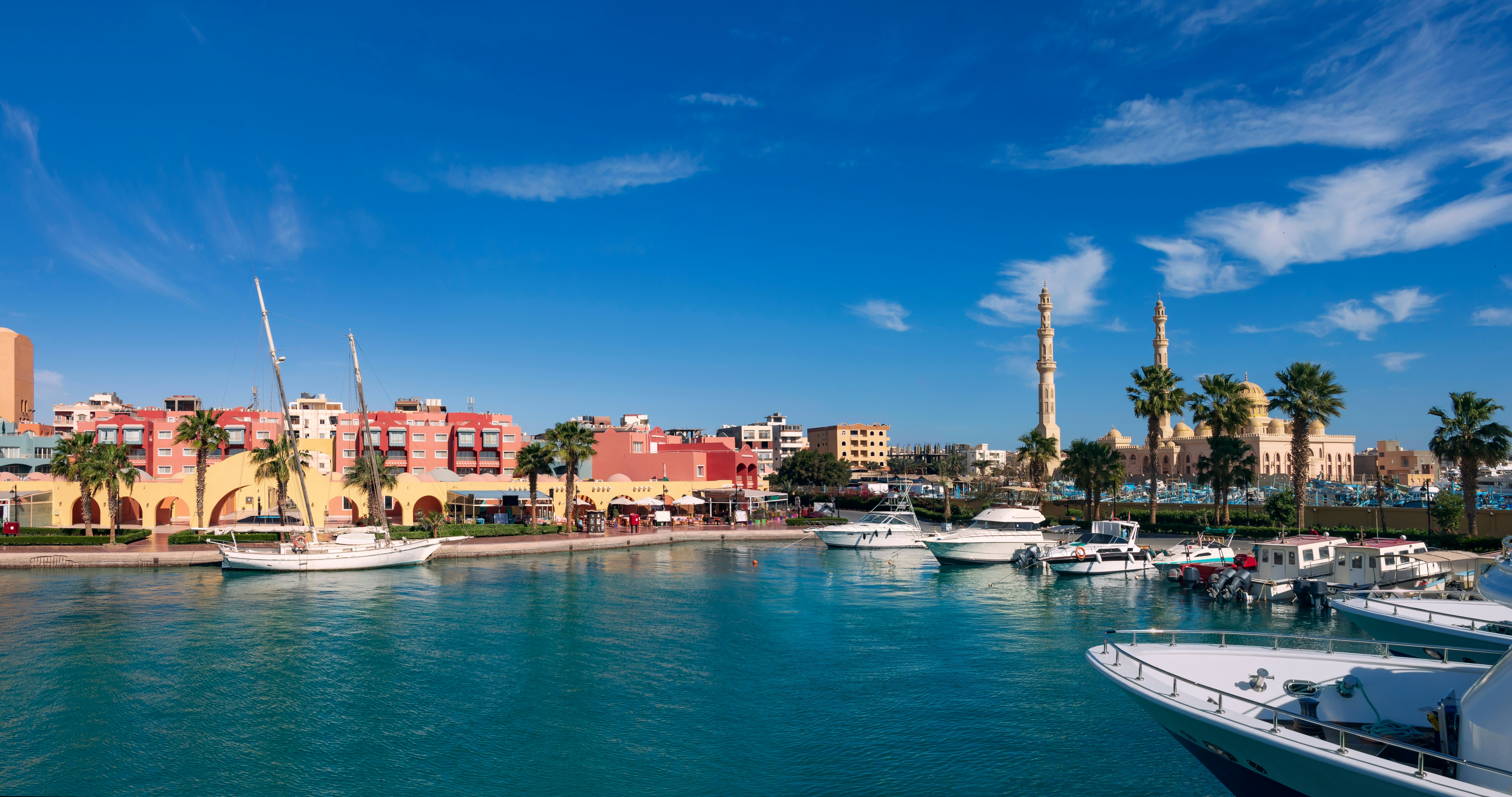 A small harbor with several docked yachts and small motorboats. A mosque with two tall minarets stands on the shore.