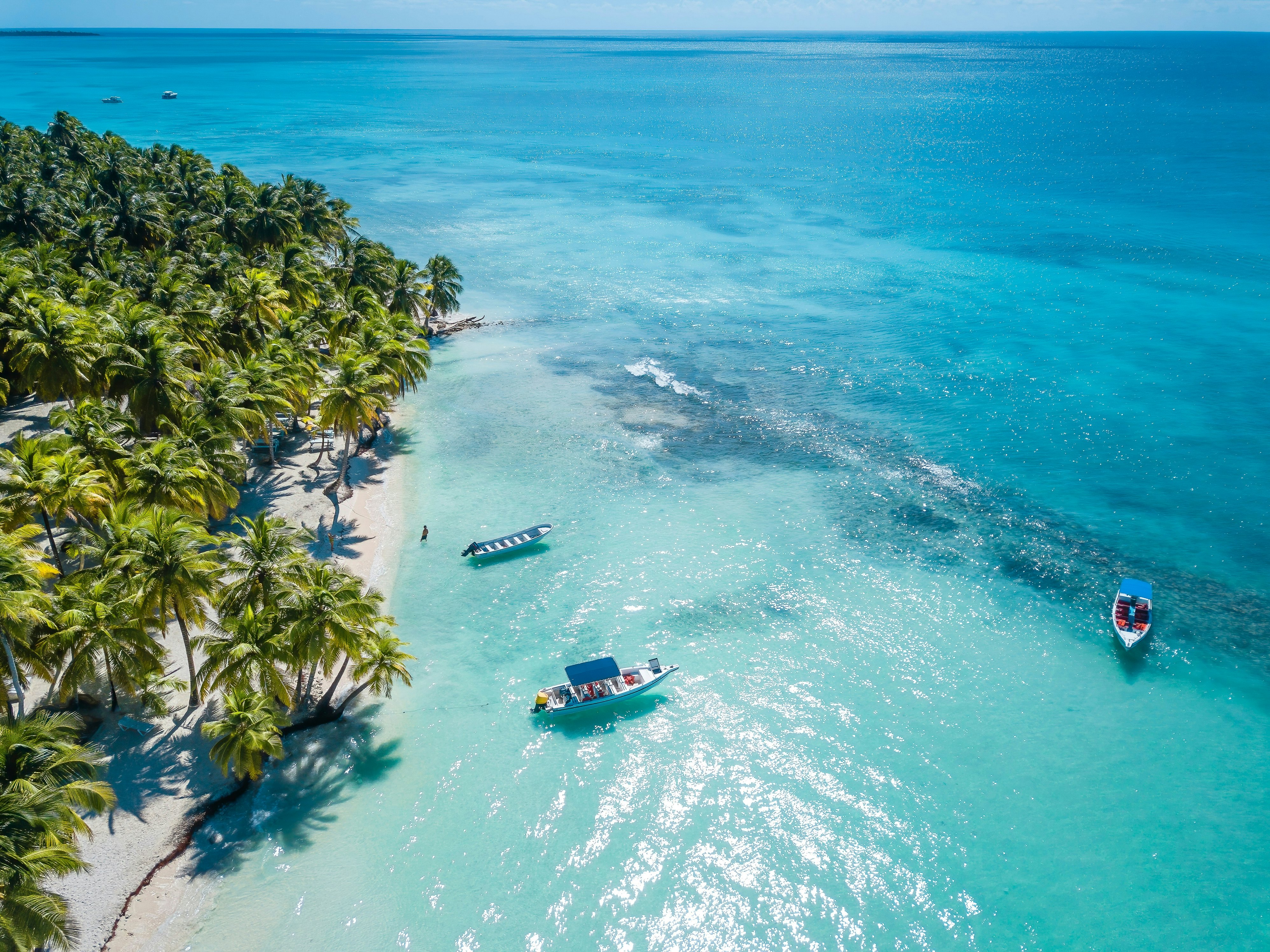 A boat floating in clear turquoise water off a sandy beach lined with palm trees.