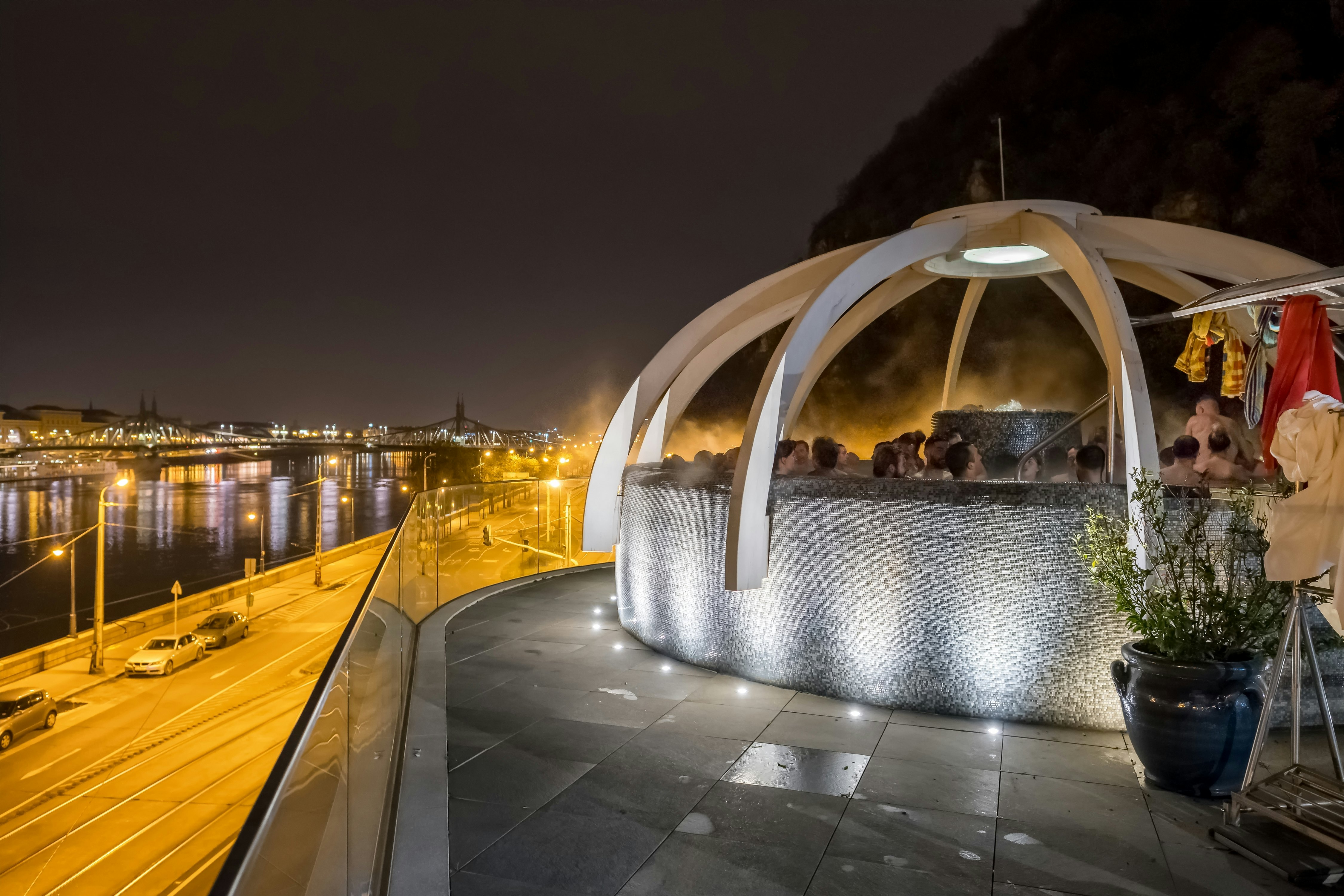 A large hot tub with lots of people in it on the roof of a building at night. A city bridge crosses a nearby river.