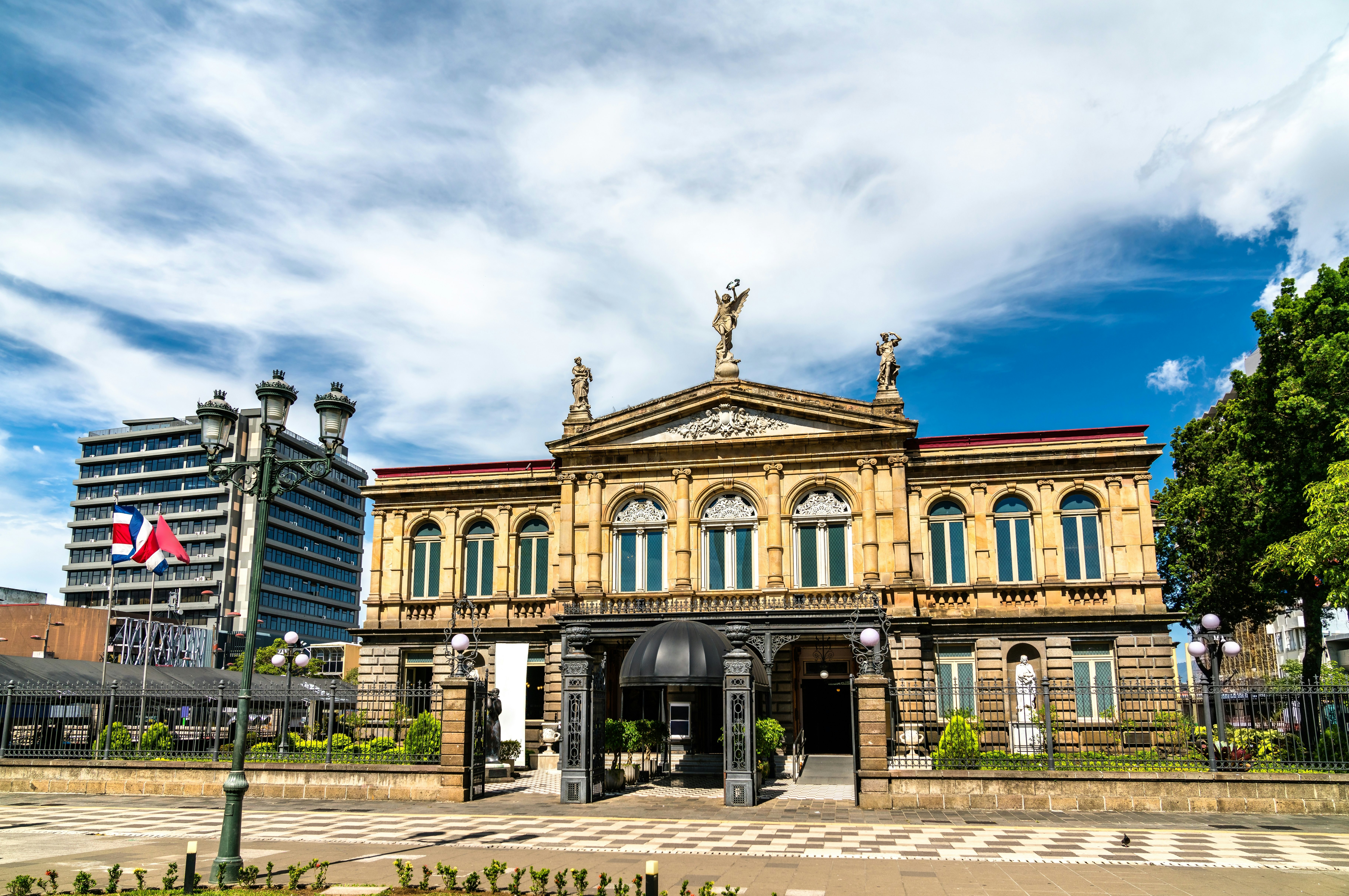 A two-story building with three statues above the three-pointed cornice and a walkway in front.