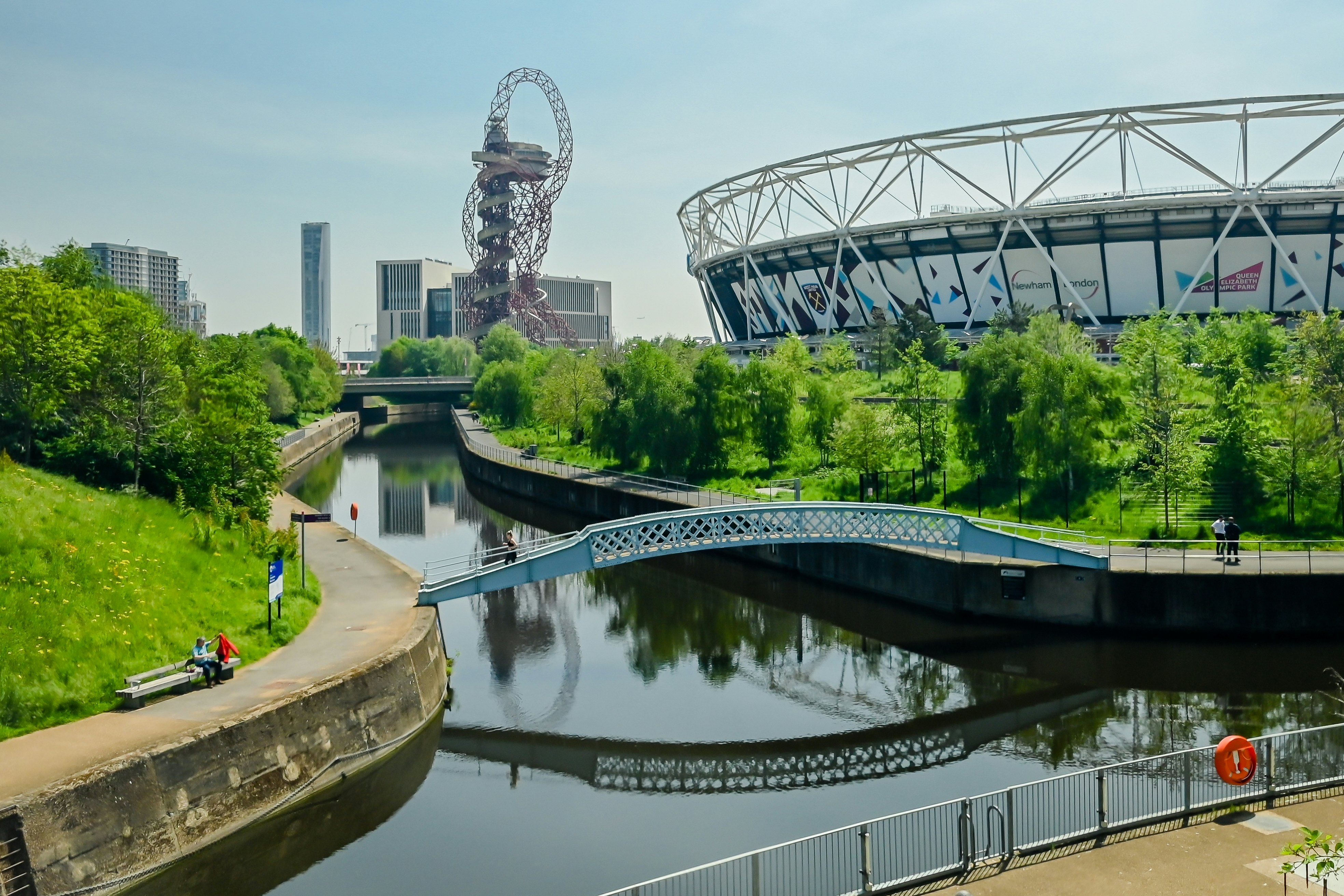 Canal bordered by cement walkways, with a stadium to the right and city buildings and an amusement ride in the distance on an overcast day.