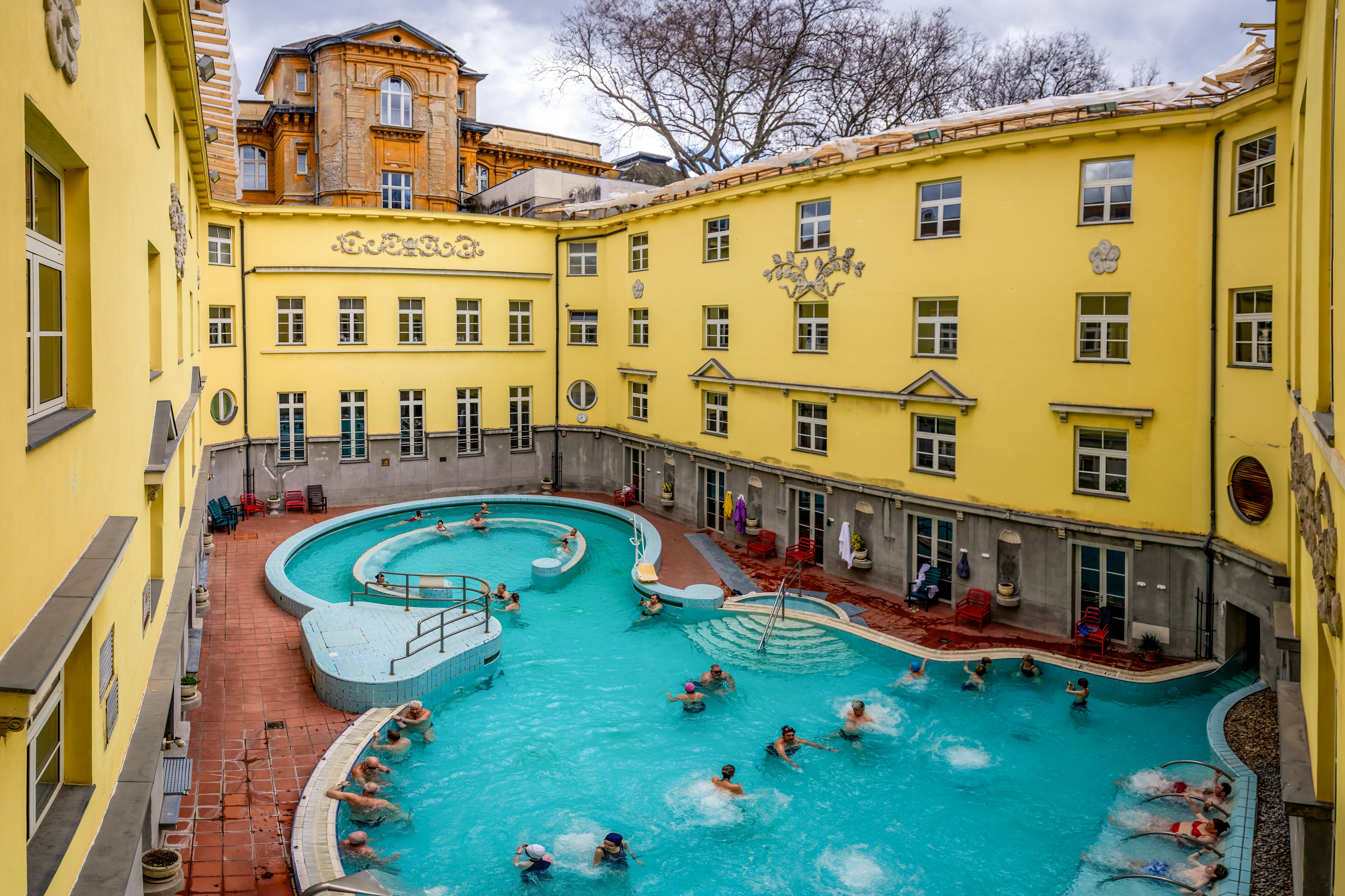 A blue pool surrounded by the walls of a yellow building. People swim, soak and relax at the sides.