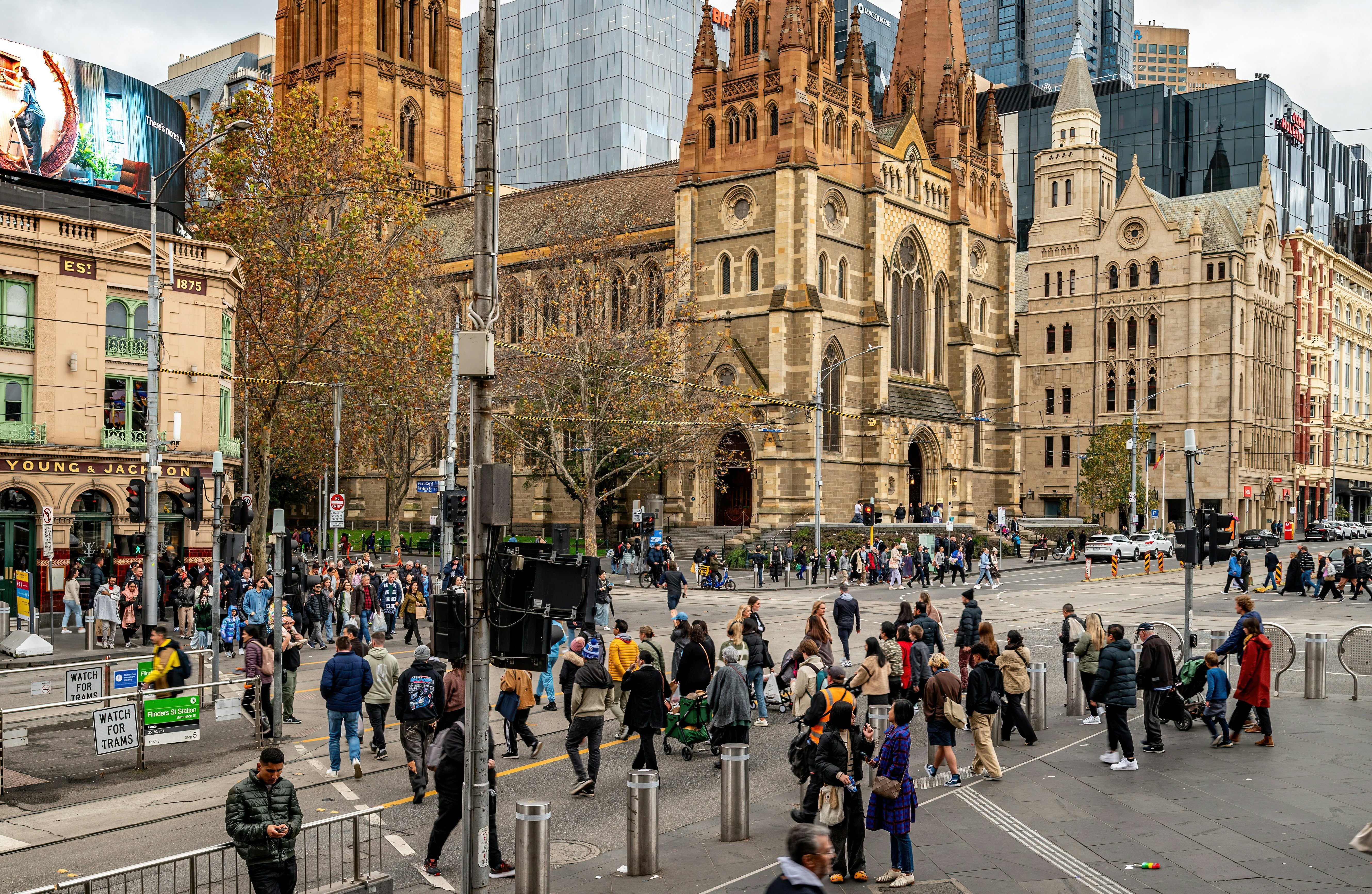 People crossing Flinders St towards Swanston St in Melbourne's CBD with St Paul's Cathedral on one corner and Young and Jackson's pub on the other