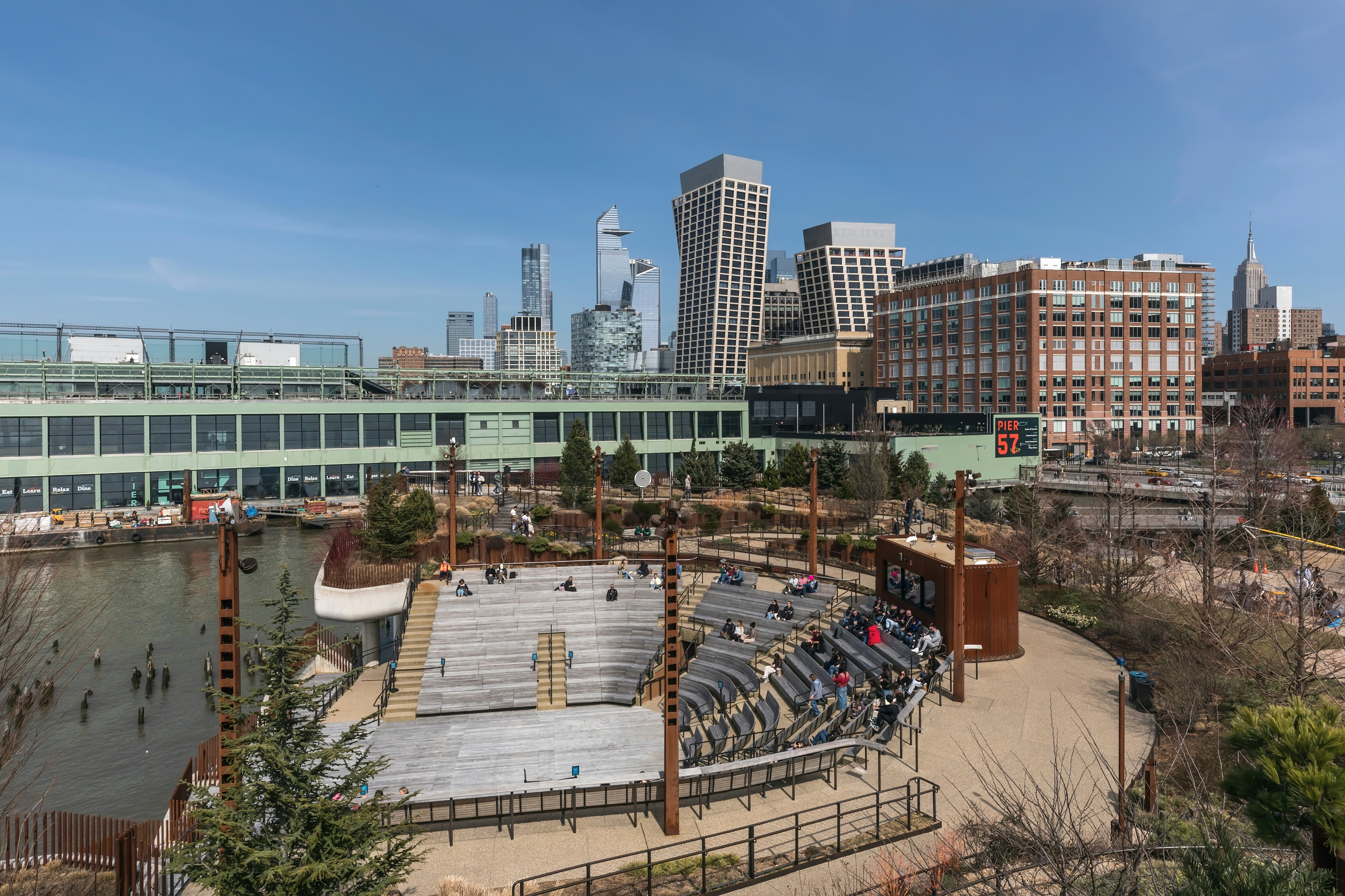 A view from above of an outdoor amphitheater at a waterside city park. Buildings of various heights are seen in the distance.