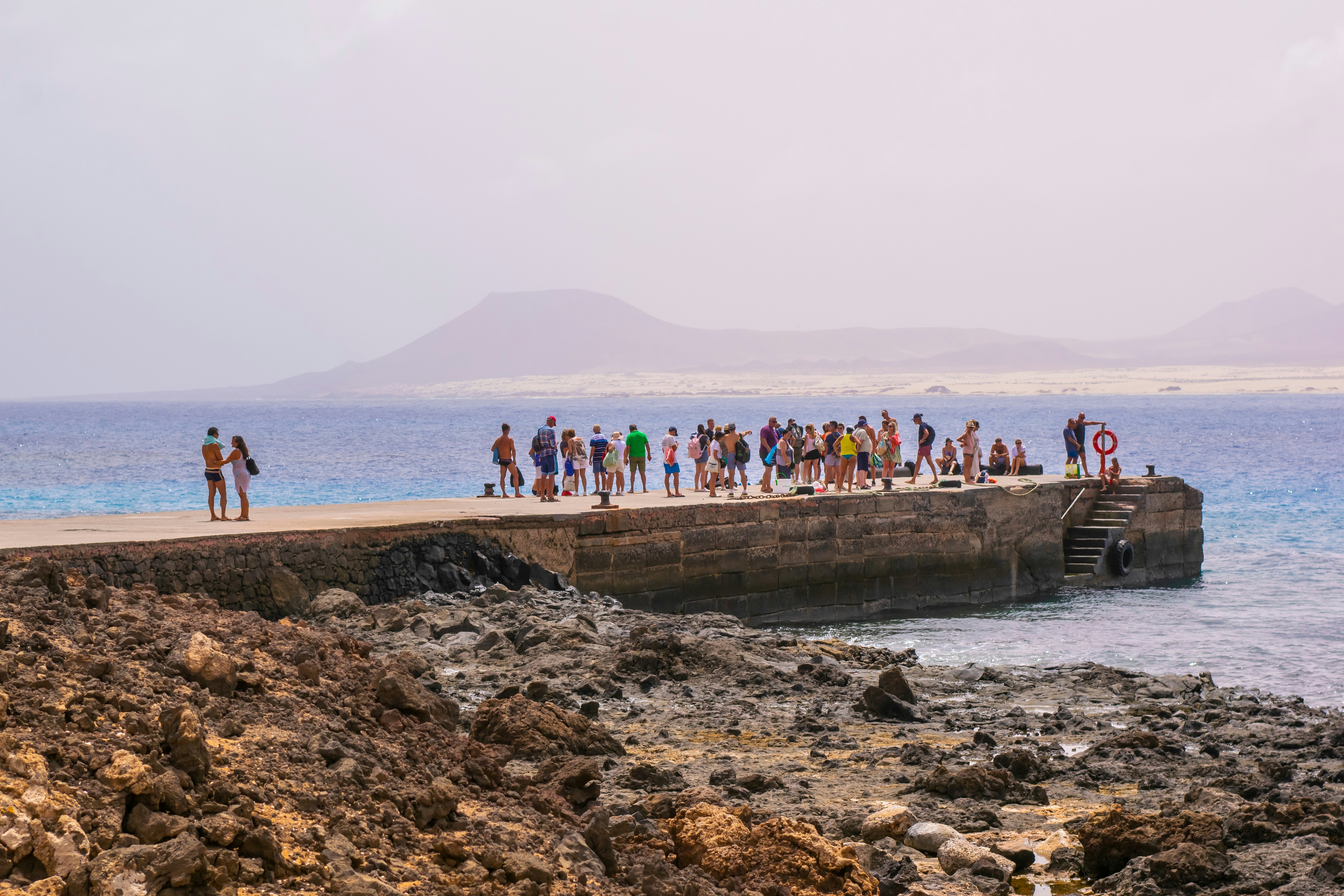People waiting for a ferry at a small harbour in the sun with beach and volcanic mountains in distance