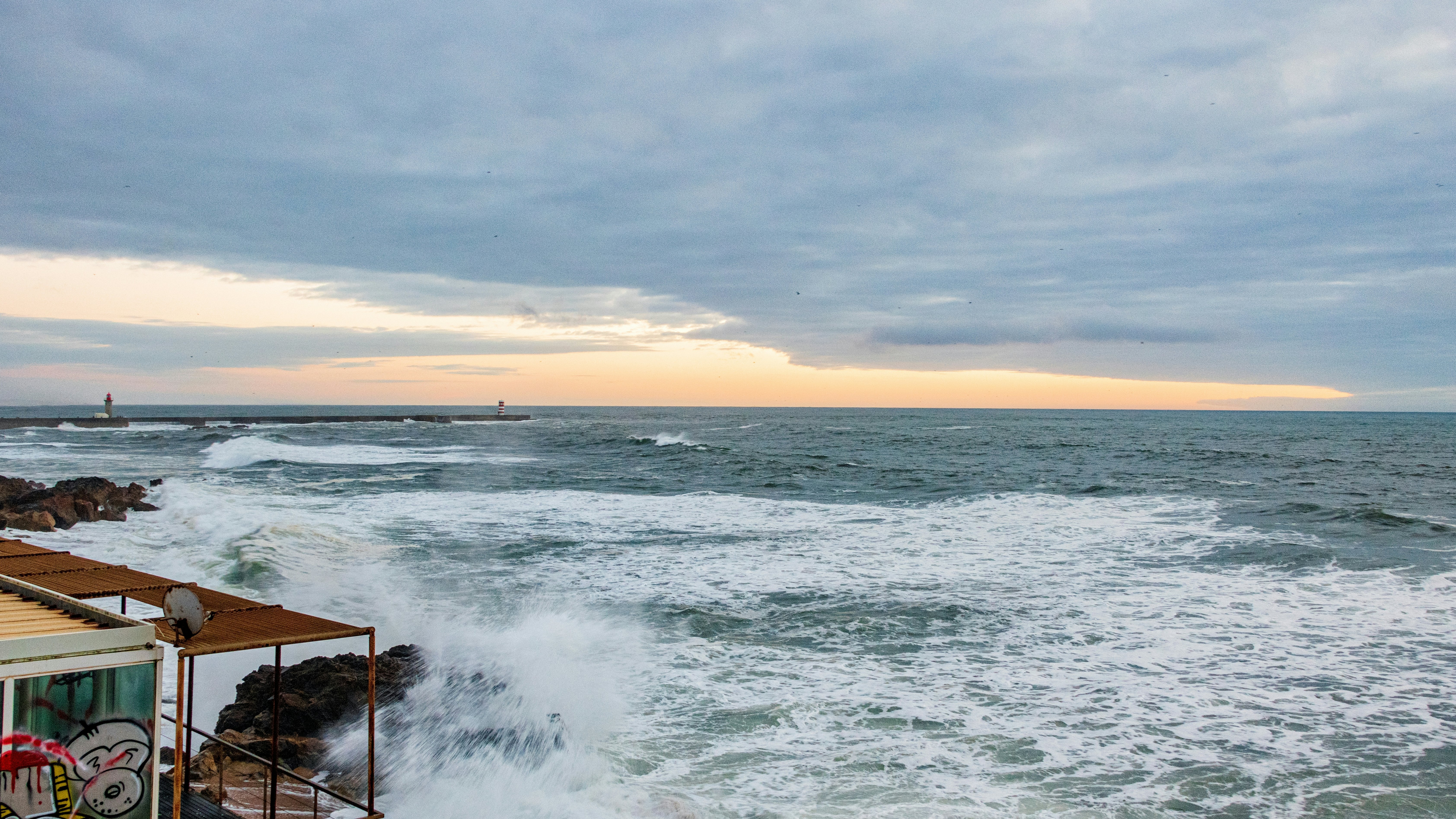 Ocean waves crashing on rocks near a building with a jetty in the far distance at sunset.