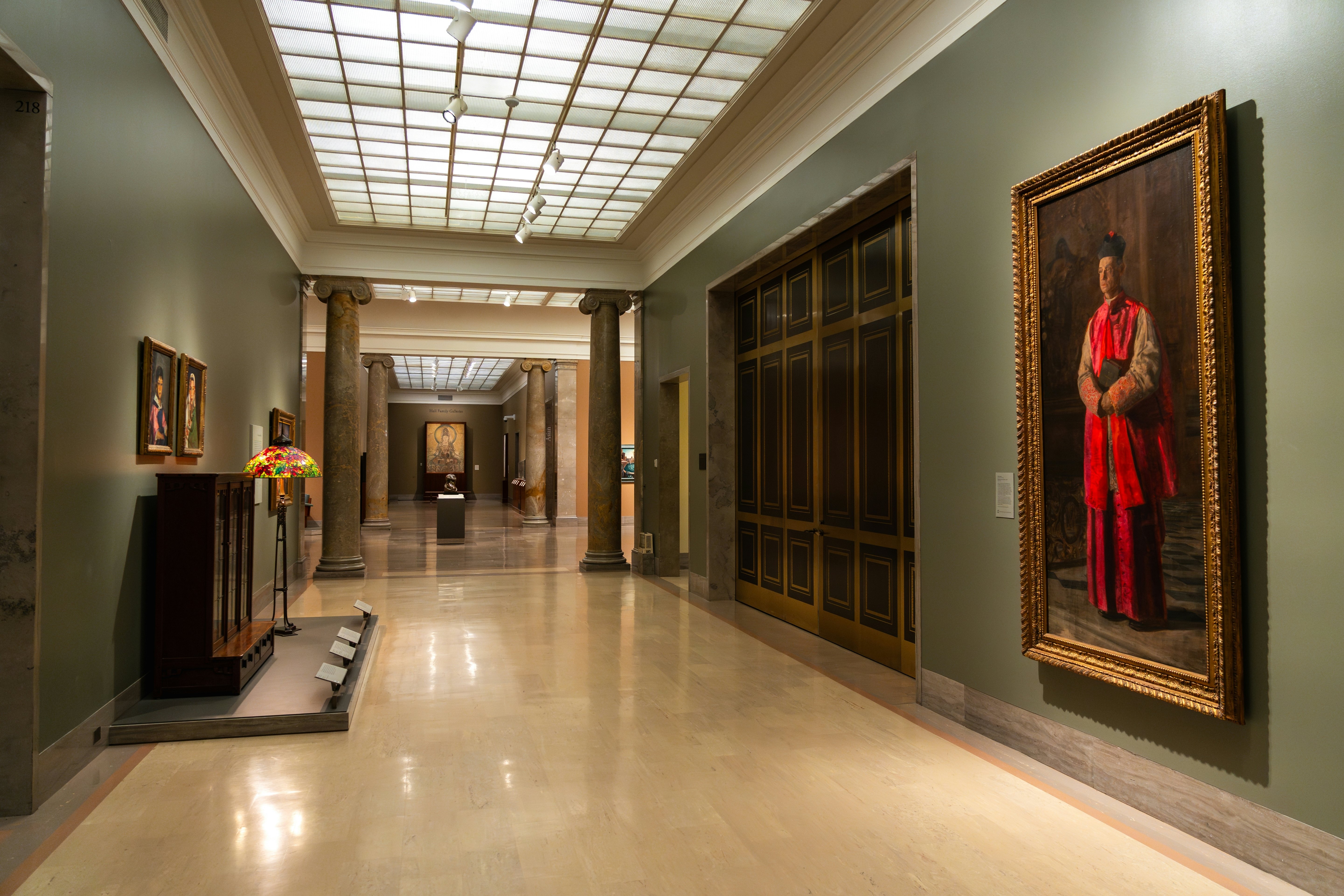 Paintings and decorative arts works are on display in a corridor of an art museum.